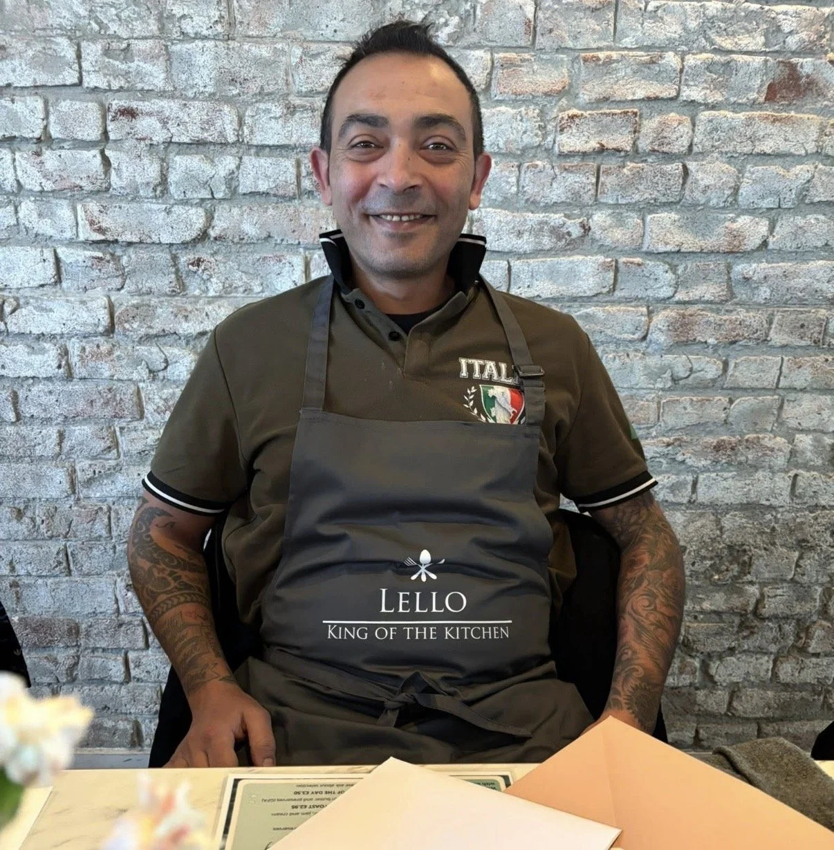A man wearing a dark apron that reads 'LELLO King of the Kitchen' sits at a table in front of a brick wall, smiling at the camera.