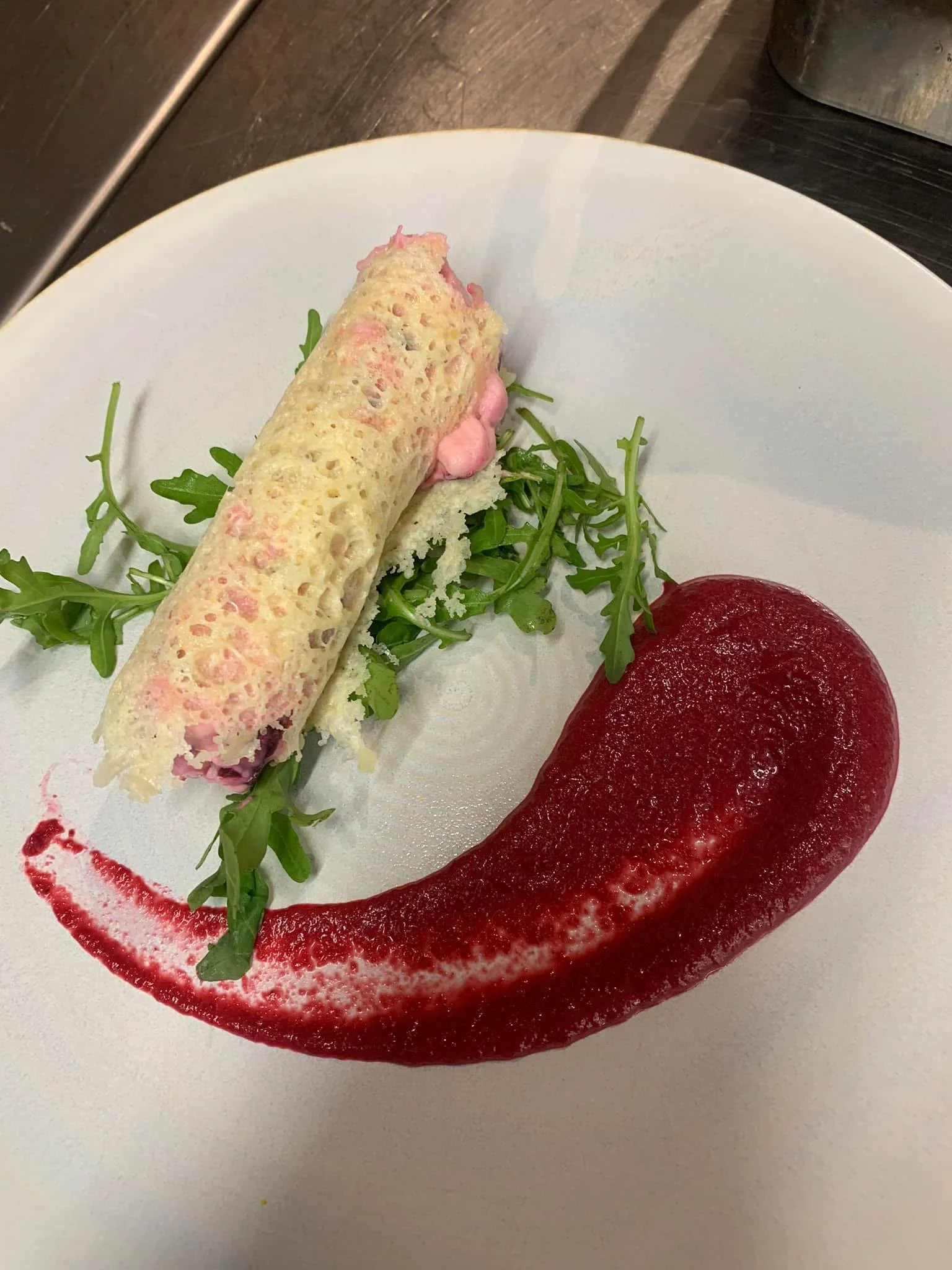 An appetizer with a rolled slice of bread or pastry with a pink filling, garnished with arugula leaves, served with a red sauce on a white plate.