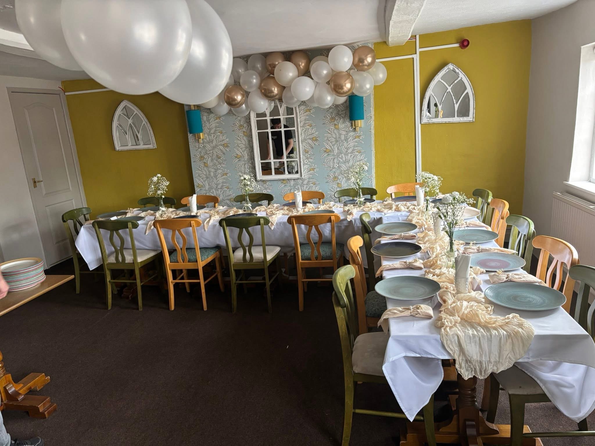 A decorated dining room with a long table set with white tablecloth, beige napkins, and plates, surrounded by colorful chairs. The wall behind has an arrangement of white, gold, and beige balloons, with a decorative mirror and wall lamps. There are small flower arrangements on the table.