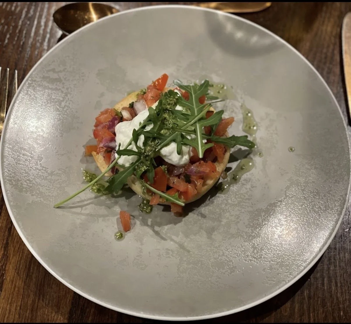 A gourmet dish with diced tomatoes, red onion, chopped herbs, topped with arugula and a dollop of cream or yogurt, drizzled with green sauce on a white plate.