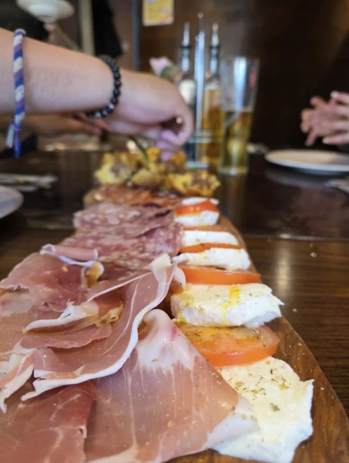 Close-up of a charcuterie board with various sliced cured meats, cheeses, and tomato slices on a wooden table, with glasses of beer and people’s hands in the background.