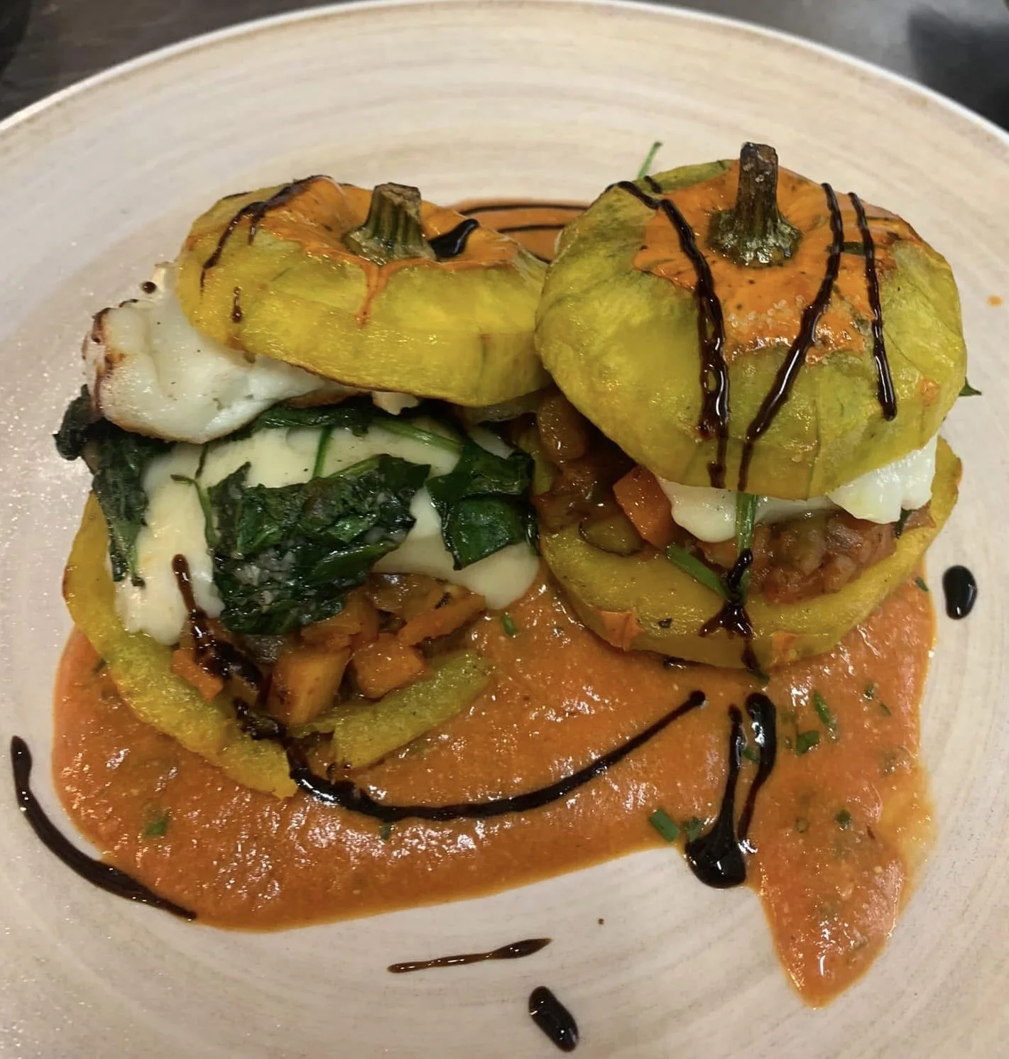 Two stuffed yellow squash on a plate with sauce and balsamic glaze drizzle, accompanied by greens and vegetables, served over a reddish-orange sauce.