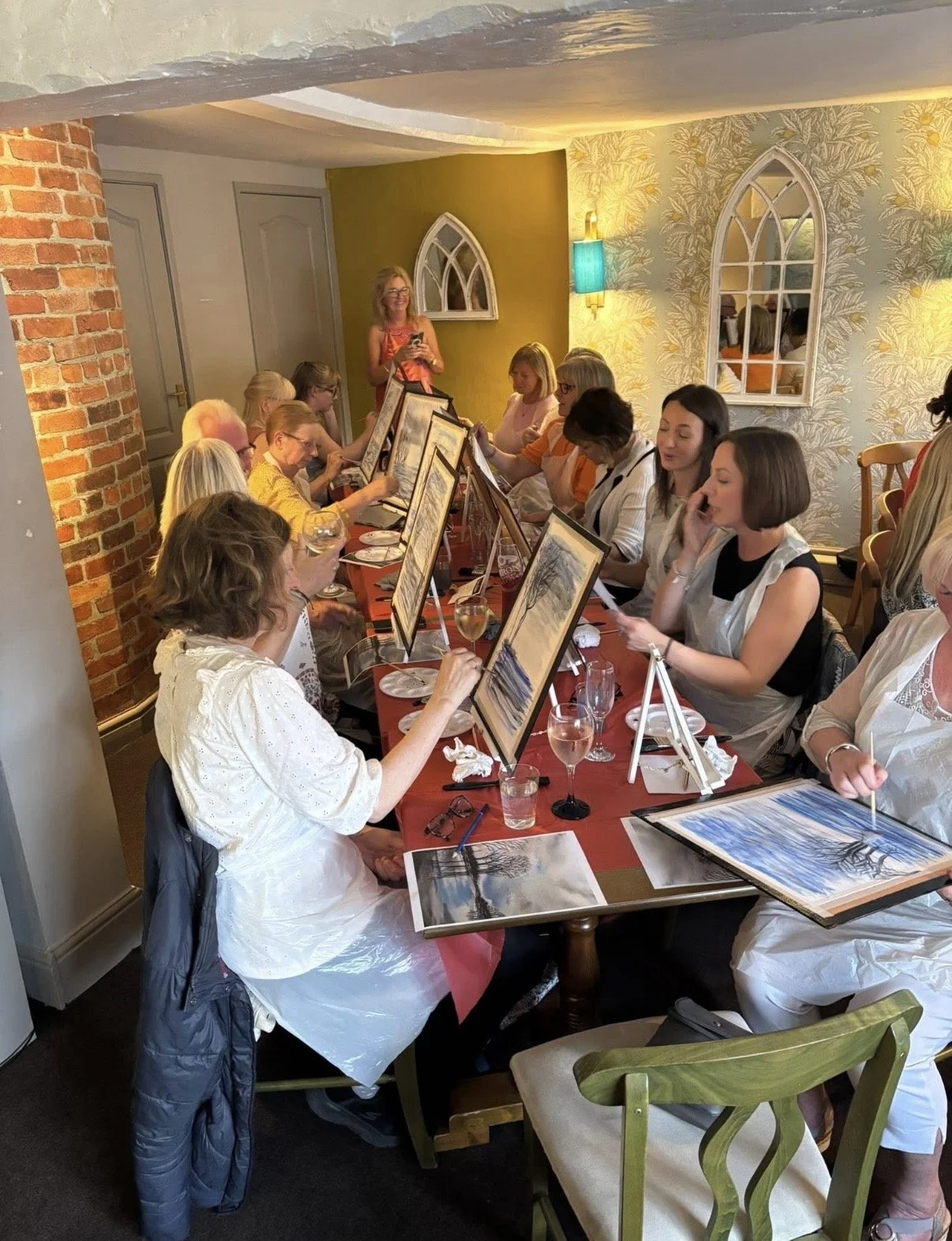 A group of women participating in a painting class, sitting at a long table with art supplies, while an Instructor demonstrates painting techniques in a cozy, decorated room.