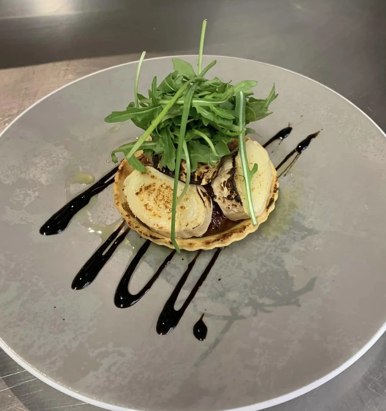 A plate with seared scallops on a tart shell, drizzled with balsamic reduction, topped with fresh greens, served on a gray plate.