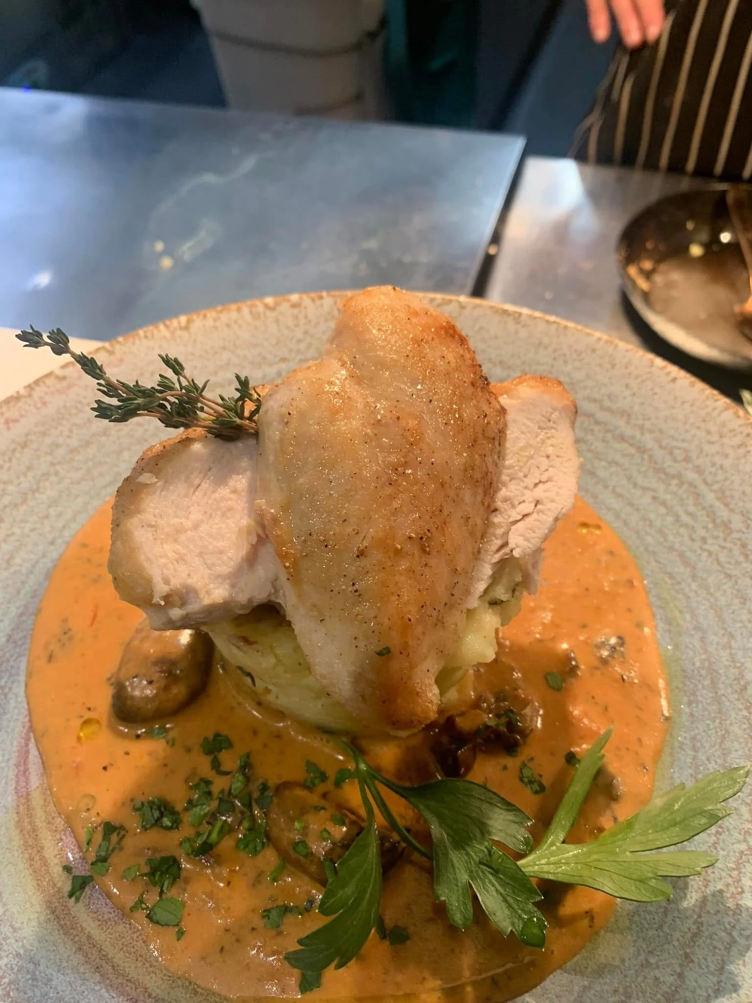 A plated dish featuring a piece of cooked chicken topped with herbs, served with a creamy sauce with mushrooms and garnished with fresh herbs and greens.