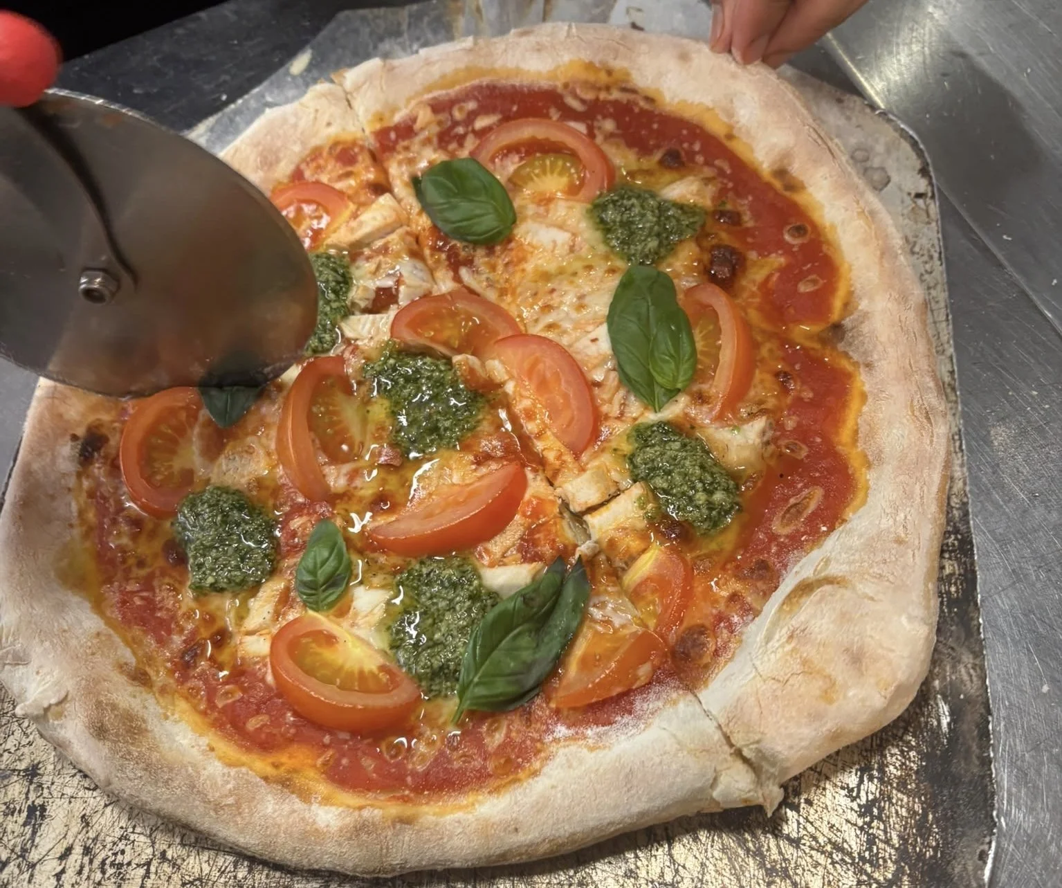 A freshly baked pizza with tomato slices, basil leaves, cheese, and green pesto on a metal tray.