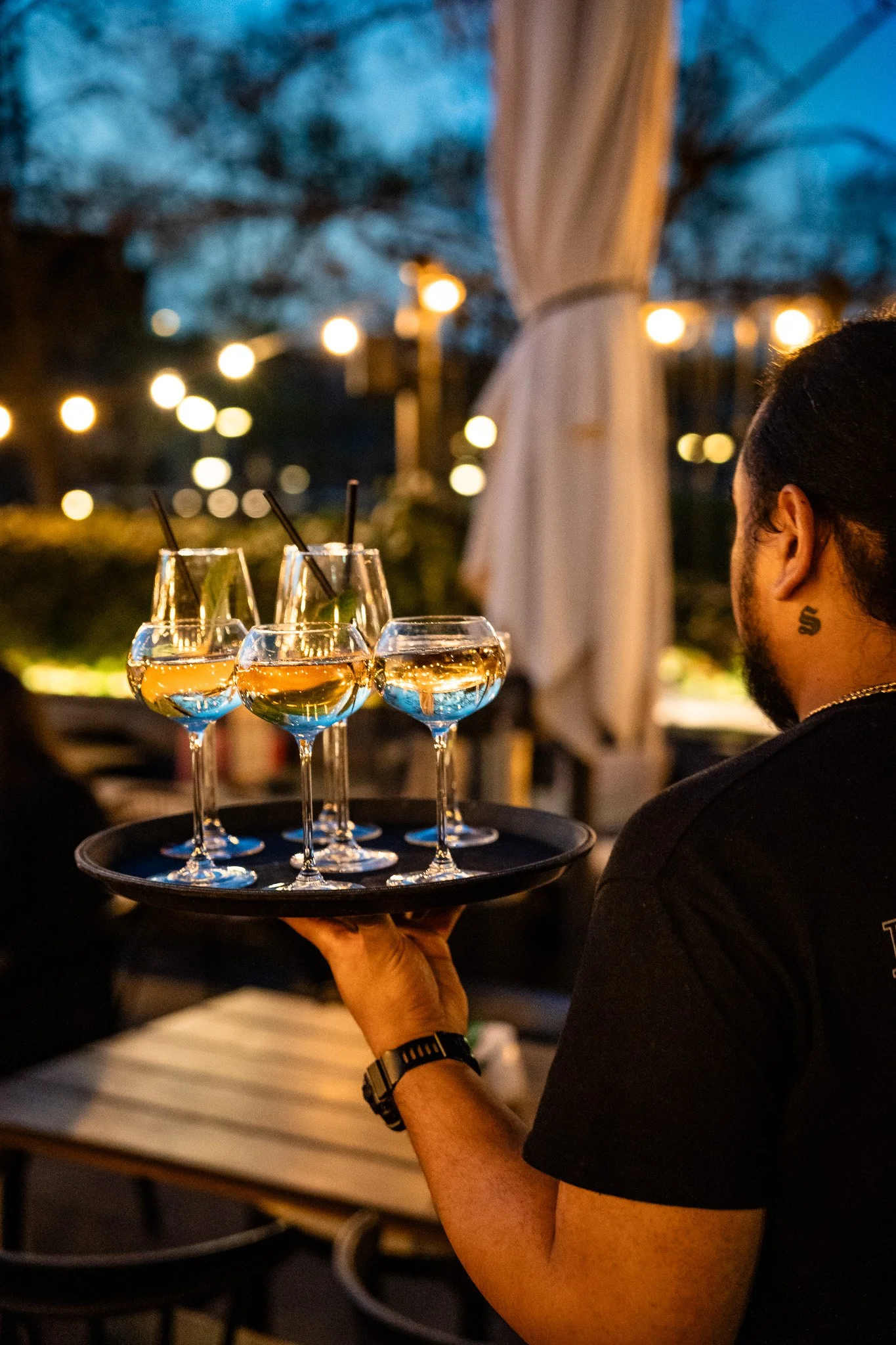 A waiter serving a tray of four glasses with drinks at an outdoor dining area during evening, with string lights and trees in the background.