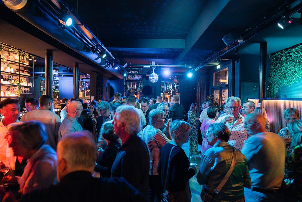 Crowd of people socializing and drinking at a bar or nightclub with colorful lighting and shelves stocked with bottles in the background.