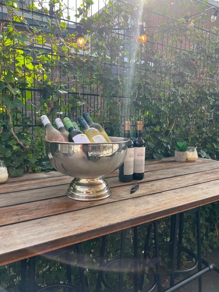 Outdoor wooden table holding several wine bottles, a large silver bowl with more bottles inside, two bottles of red wine, a small potted plant, and candles in glass holders, set against a backdrop of green ivy on a fence.