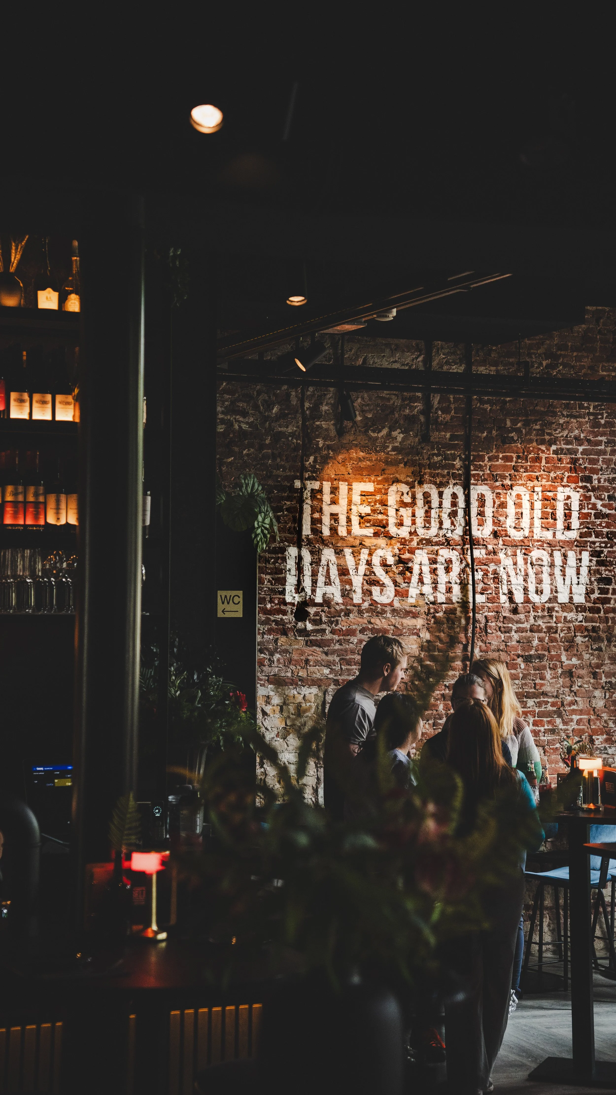 Inside a dimly lit restaurant with an exposed brick wall that has a large mural reading 'The Good Old Days Are Now'. Several people are dining. There is a shelf with bottles and glasses on the left and a small sign with 'WC' written on it, with an arrow pointing left.