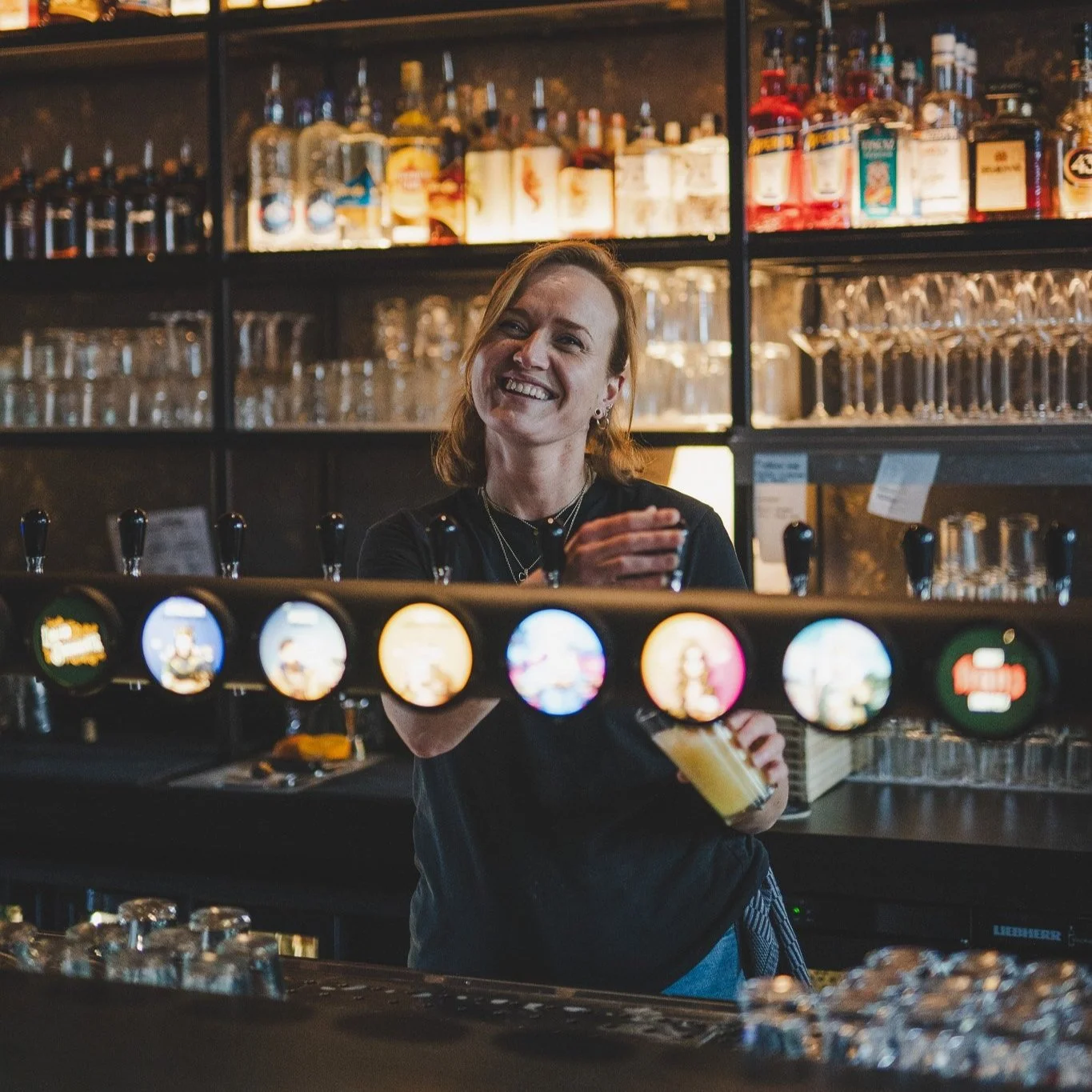 A woman bartender smiling behind a bar with beer taps in a dimly lit pub or brewery, with liquor bottles and glassware on shelves in the background.