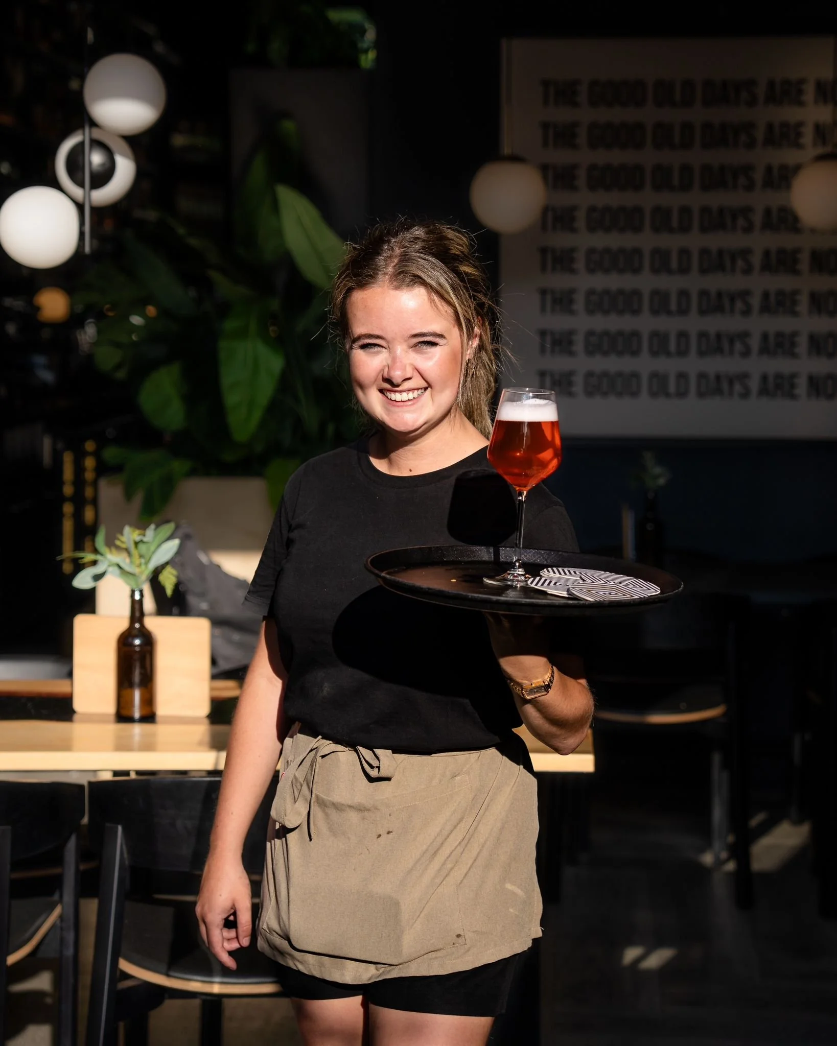 A young woman smiling, holding a tray with a glass of reddish drink, standing in a dimly lit indoor setting, with decorative lighting and plants in the background.