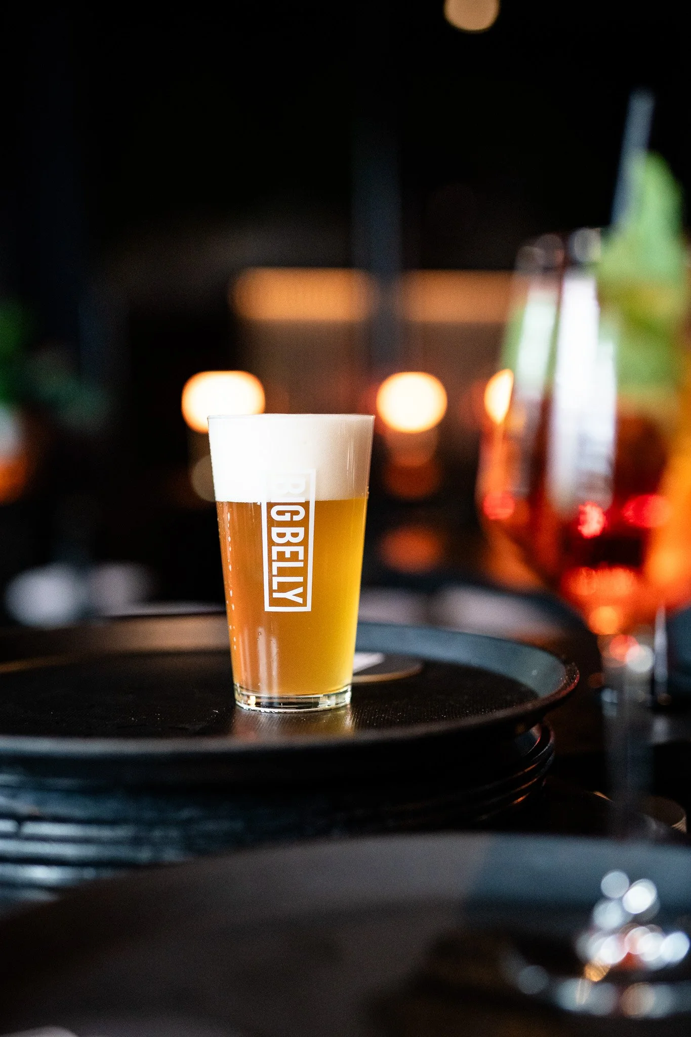 A tall glass of beer with a foam head placed on a black tray, with blurred warm lights in the background.