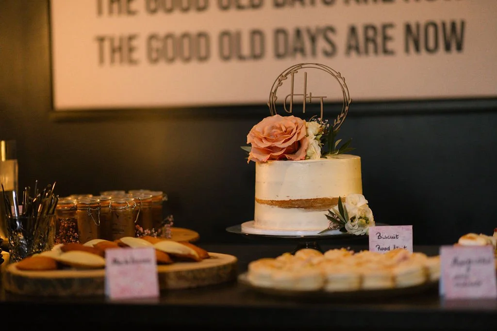 A decorated wedding cake with pink and white flowers and a circular topper, surrounded by cookies and jars on a table.