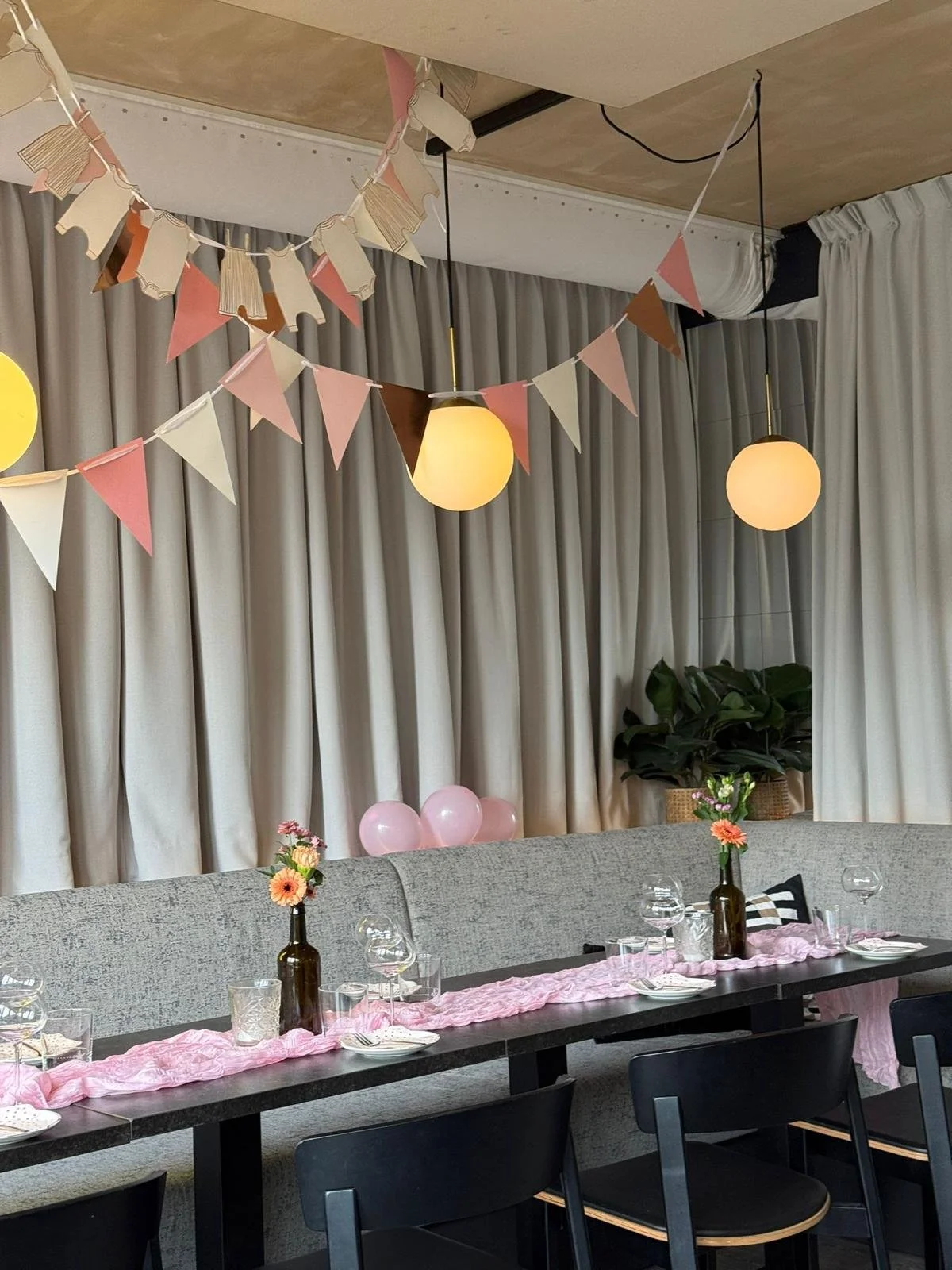 Decorated indoor space with hanging pink, cream, and brown bunting, round pendant lights, balloon decorations, a long table with pink table runner, vases with flowers, and table settings, with closed curtains and a potted plant in the background.