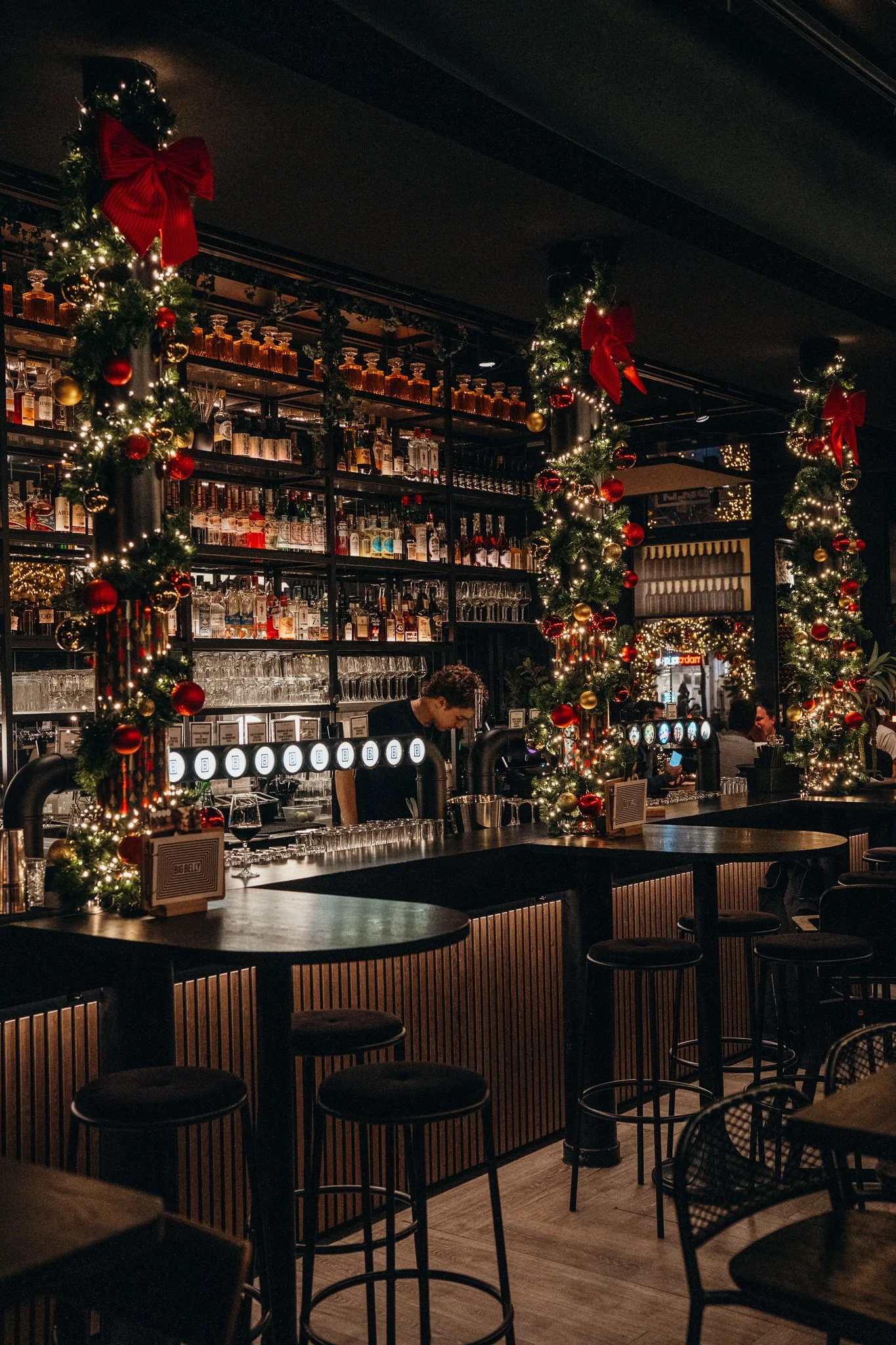 A bar decorated for Christmas with garlands, red and gold ornaments, and red bows. The bar has various bottles of alcohol and glasses on shelves behind it. The place is dimly lit with warm lighting, and there are stools and tables in the foreground.