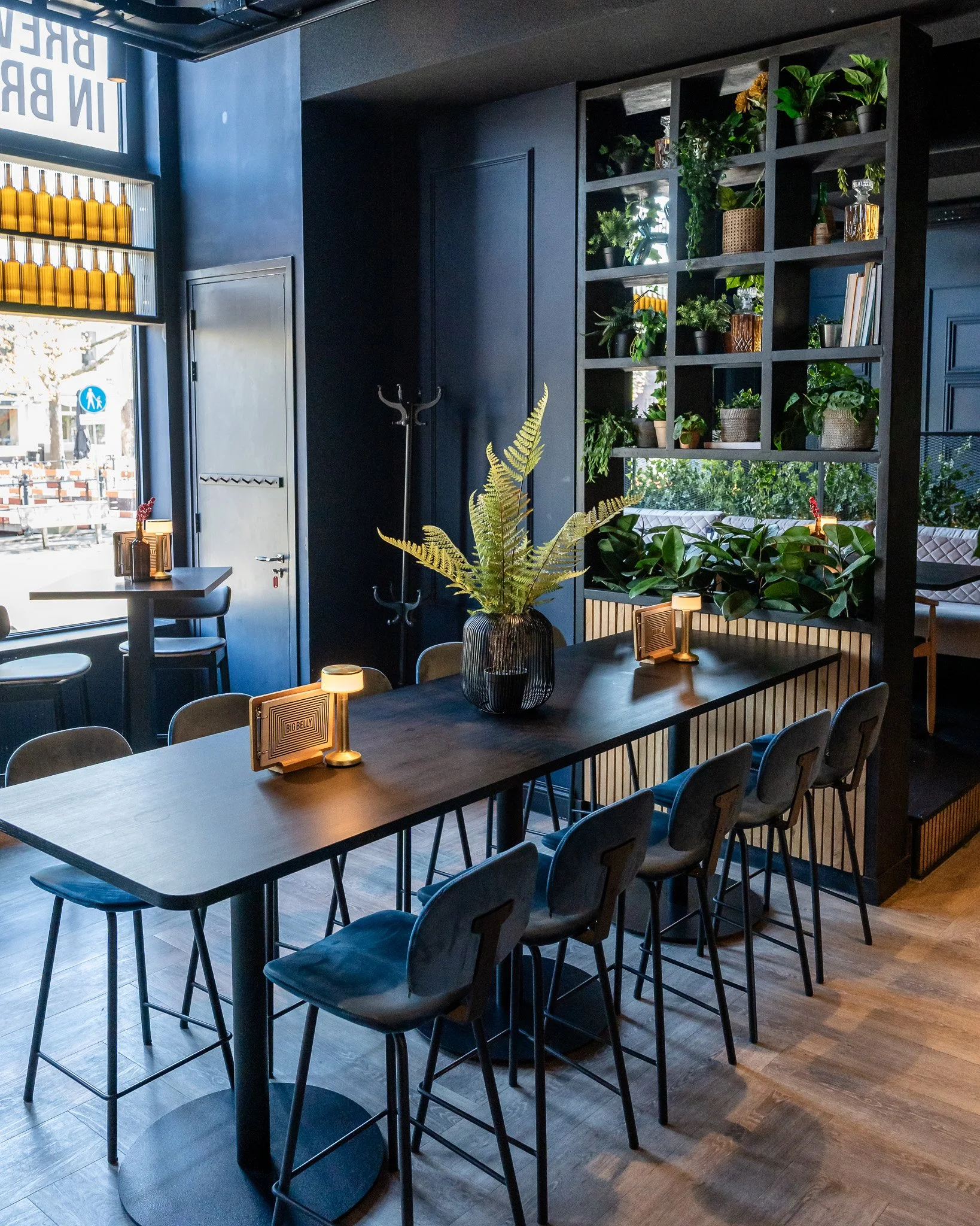 Interior of a cozy cafe with a wooden table, chairs, potted plants, and a wall shelf filled with greenery, with windows letting in natural light.