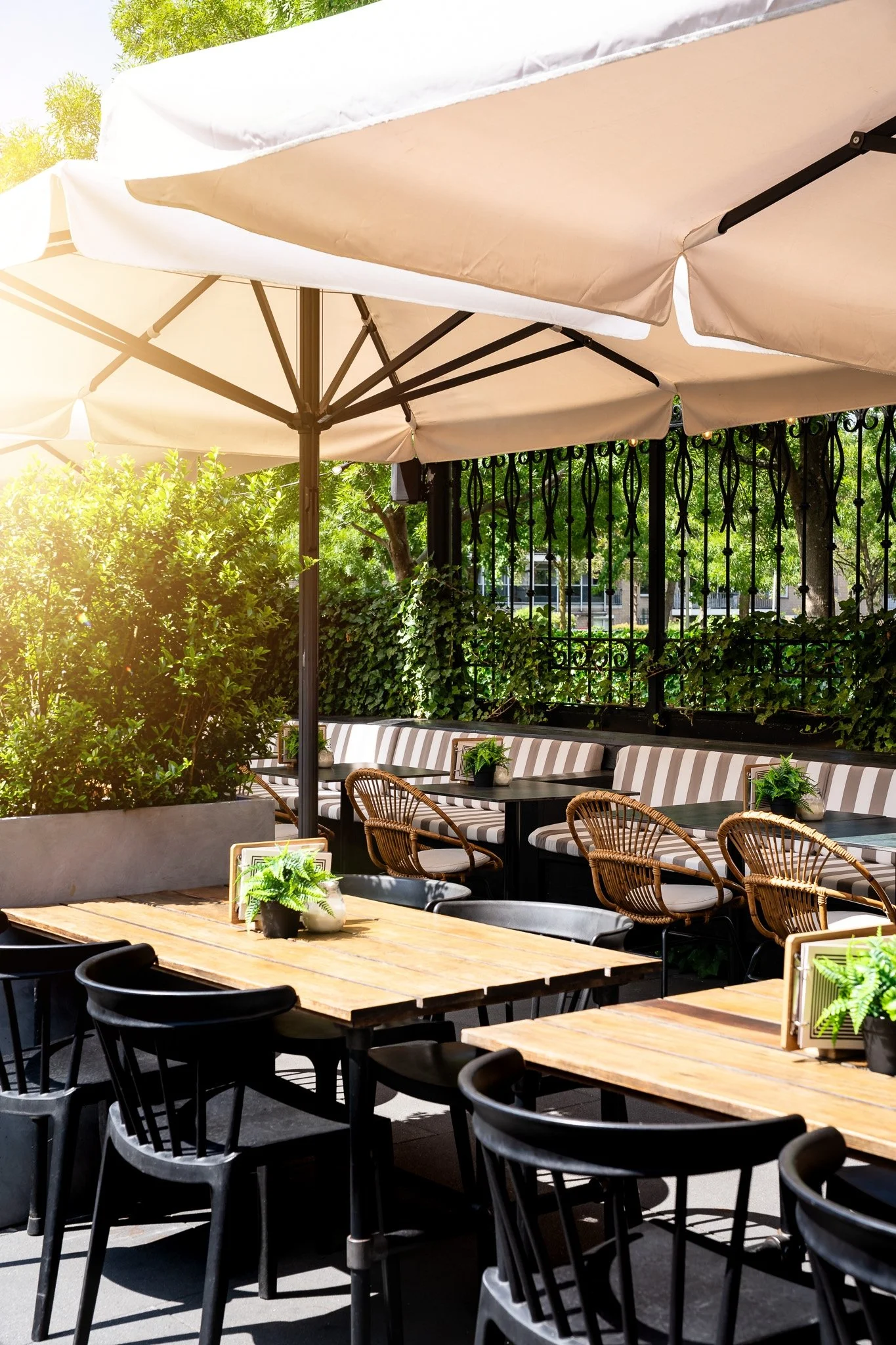 Outdoor patio seating area with black chairs, wooden tables, potted plants, striped cushioned benches, large beige umbrellas, and green foliage in the background.