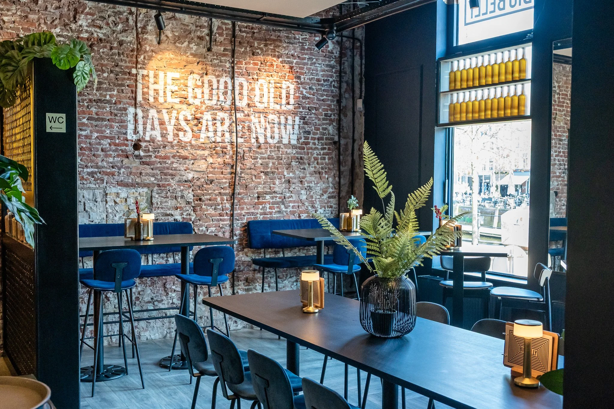 Interior of a modern cafe or restaurant with brick walls, blue chairs, a large wooden table with a black wire vase containing green fern leaves, small table lamps, and a bright window showing outdoor seating.