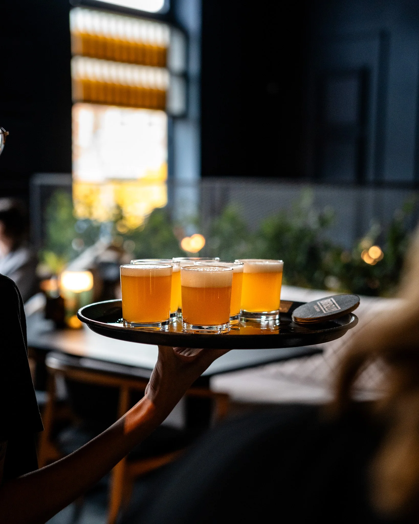 A person holding a black tray with four glasses of beer, set against a blurred background of a modern interior with dim lighting.