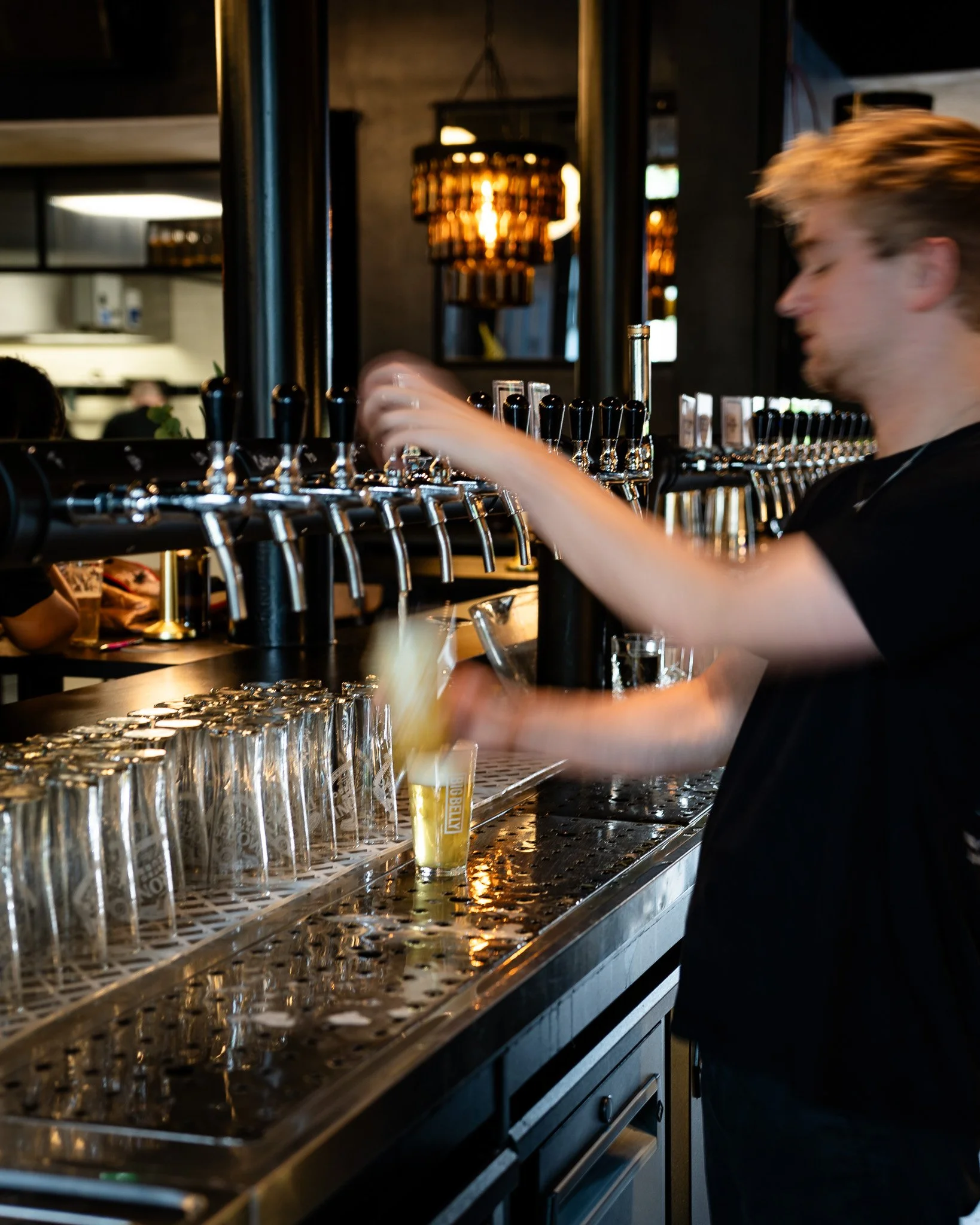 A person pouring a beer from a tap at a bar, with multiple taps and empty glasses on the counter, in a dimly lit setting.