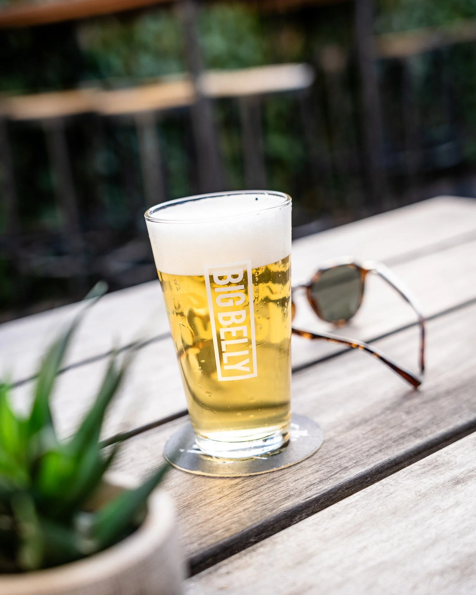 A clear glass of beer on a wooden table outdoors with a pair of sunglasses and green plant leaves nearby.