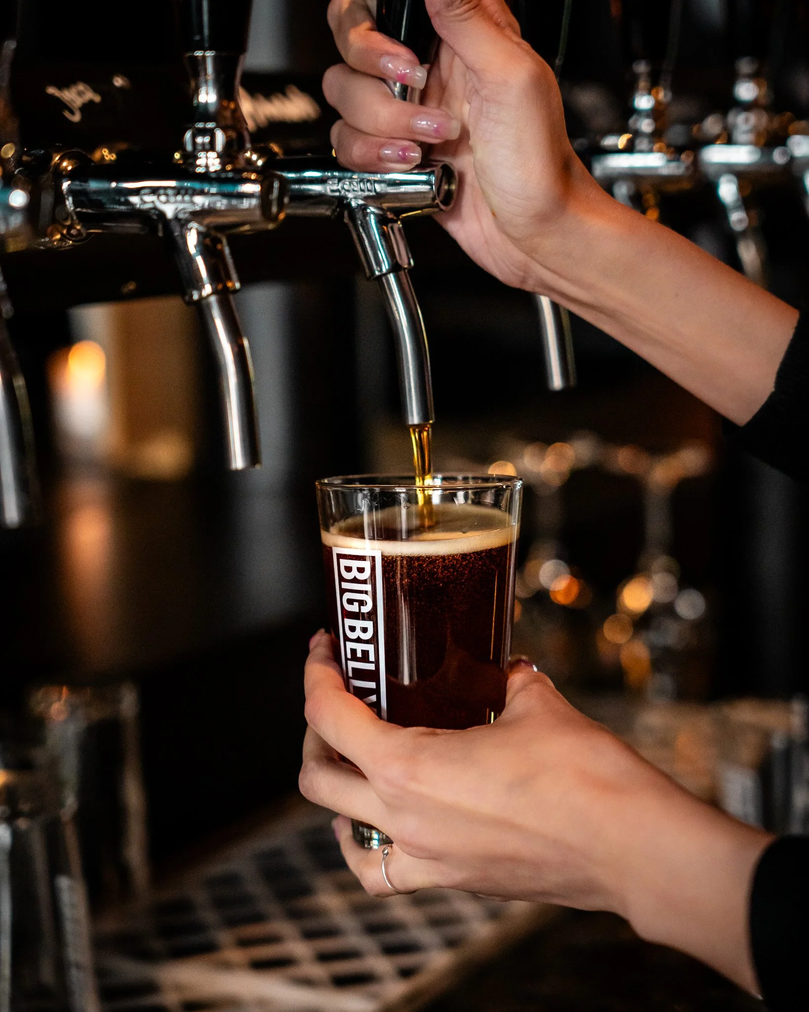 Person pouring beer from tap into glass labeled 'Big Belly' at bar.