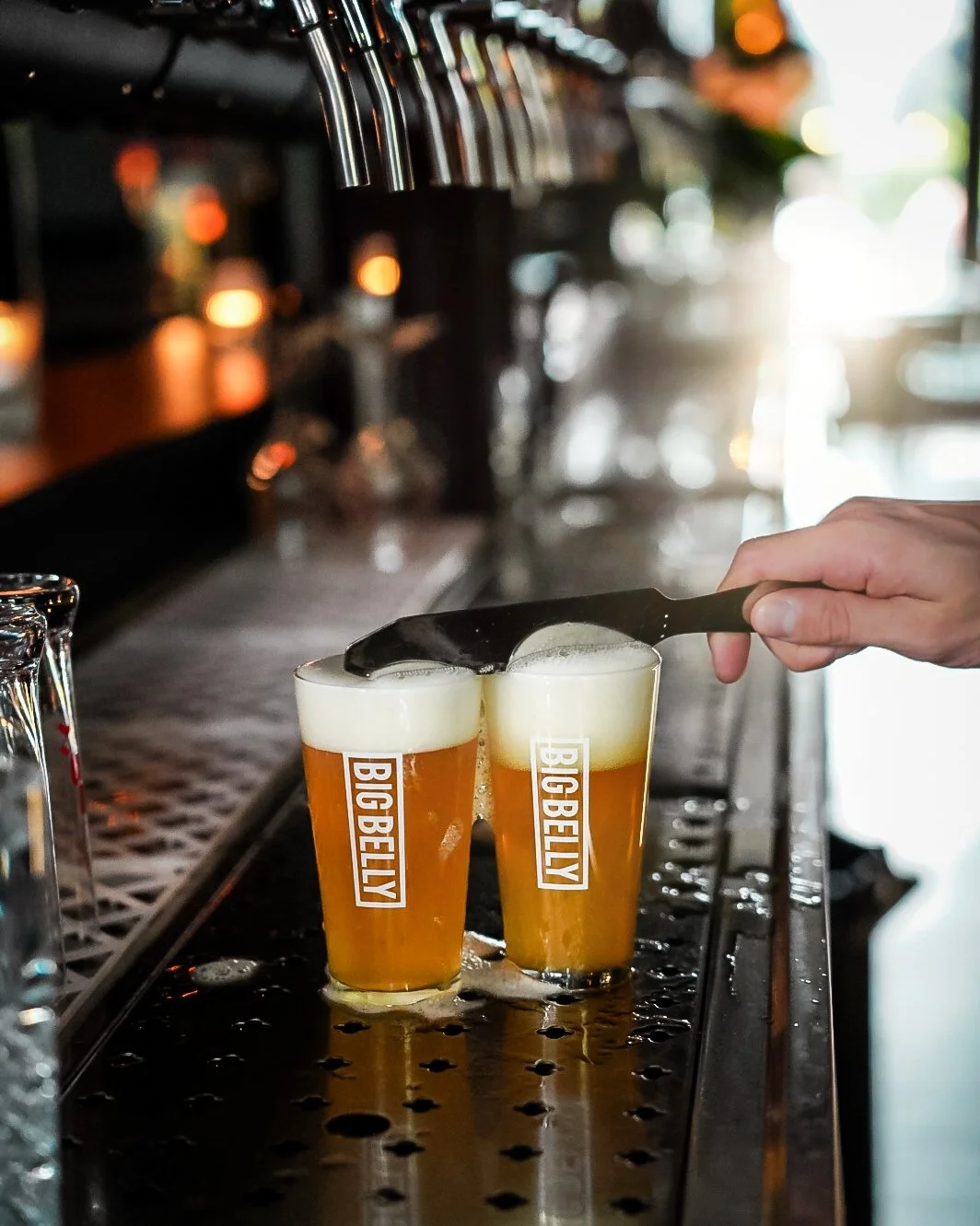 Beer glasses labeled 'BIG BELLY' on a bar counter with a hand holding a scraper to remove foam.