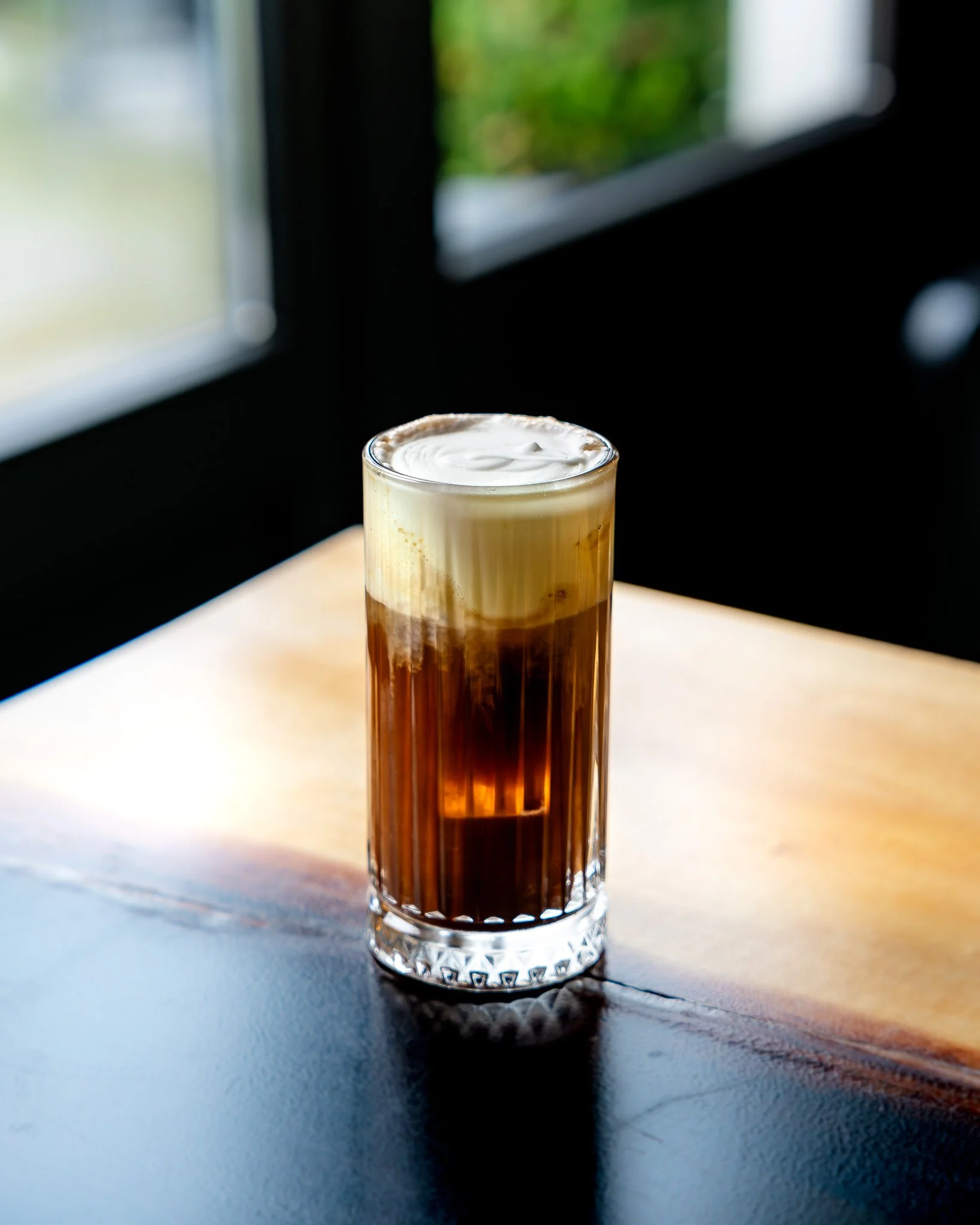 A tall glass with layered coffee beverage, topped with whipped cream, on a dark textured surface near a window with green outdoor scenery.