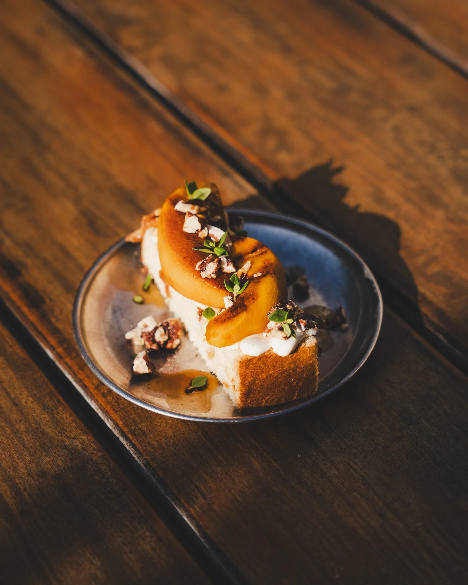 A dessert on a dark plate features sliced toasted bread topped with caramelized peaches, garnished with fresh herbs, crushed nuts, and drizzled with syrup, set on a wooden surface.