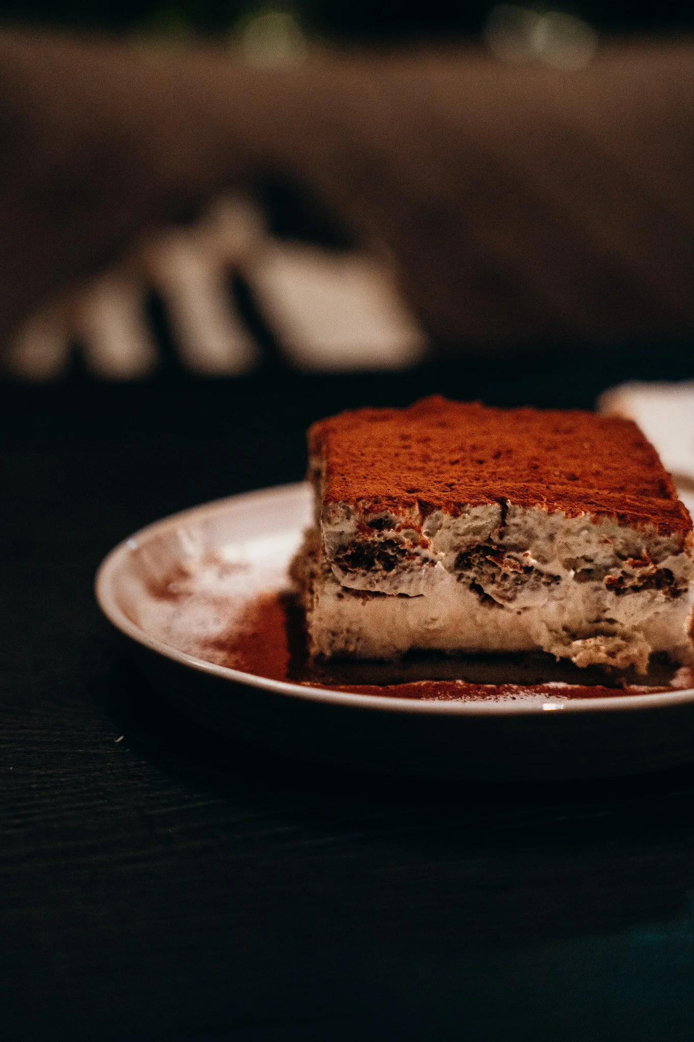 A slice of cookies and cream ice cream cake covered with cocoa powder on top, placed on a white plate with a silver spoon beside it, on a dark surface.