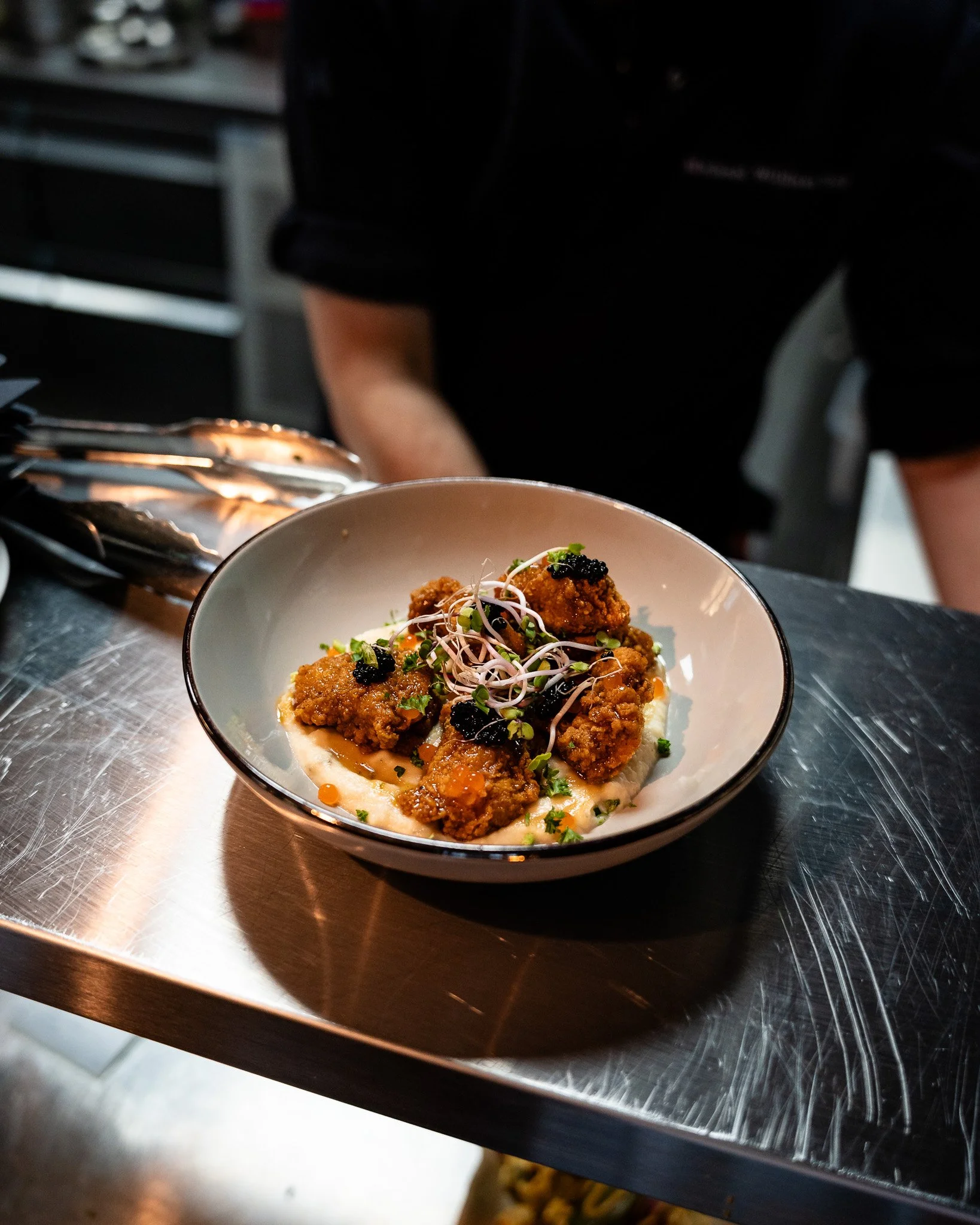A close-up of a bowl of fried chicken pieces garnished with microgreens, served with caviar, on a metal countertop in a professional kitchen.
