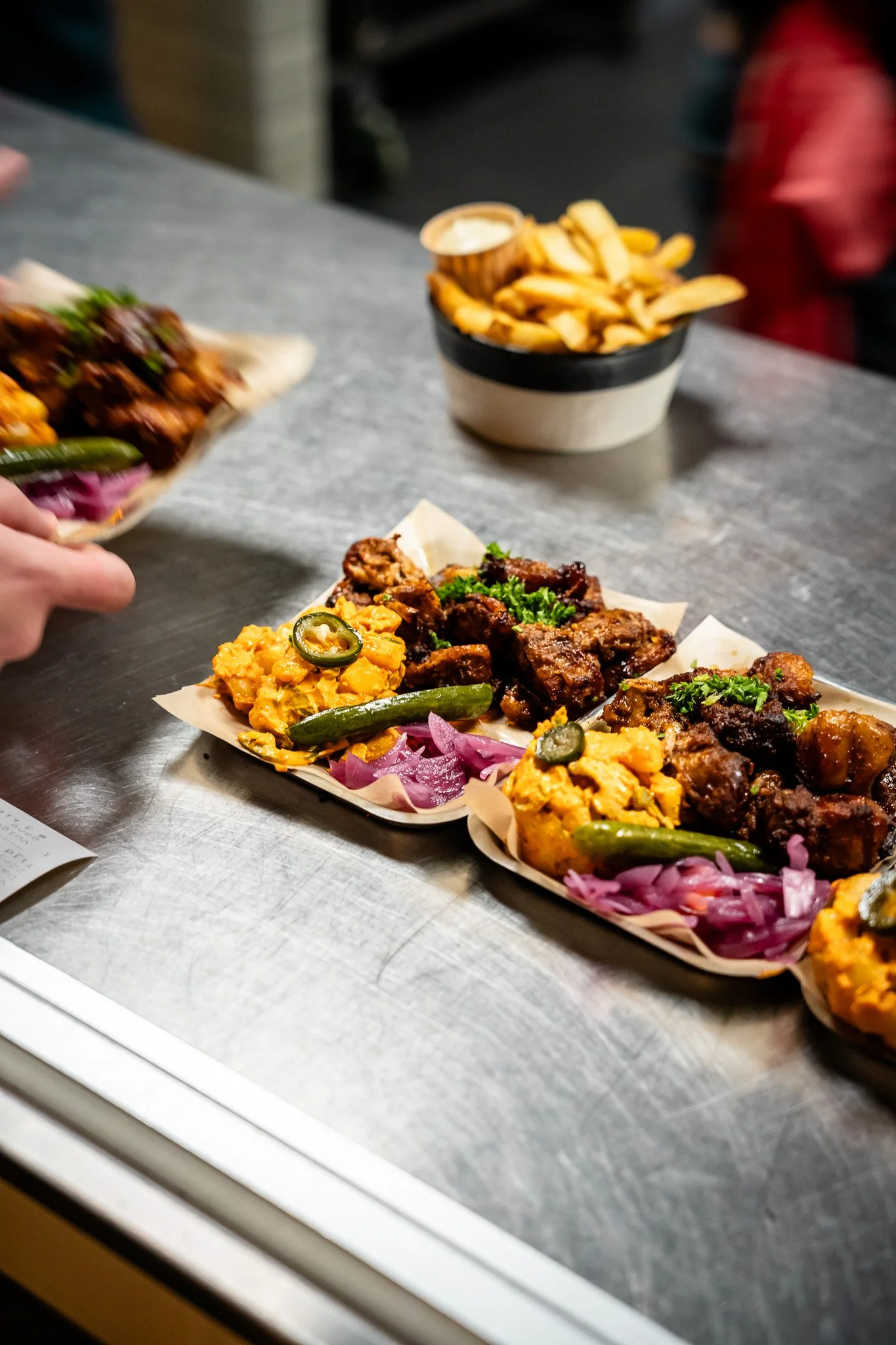 A close-up of a food tray with fried chicken, mac and cheese, and pickles, and a bowl of French fries with dipping sauce in the background.