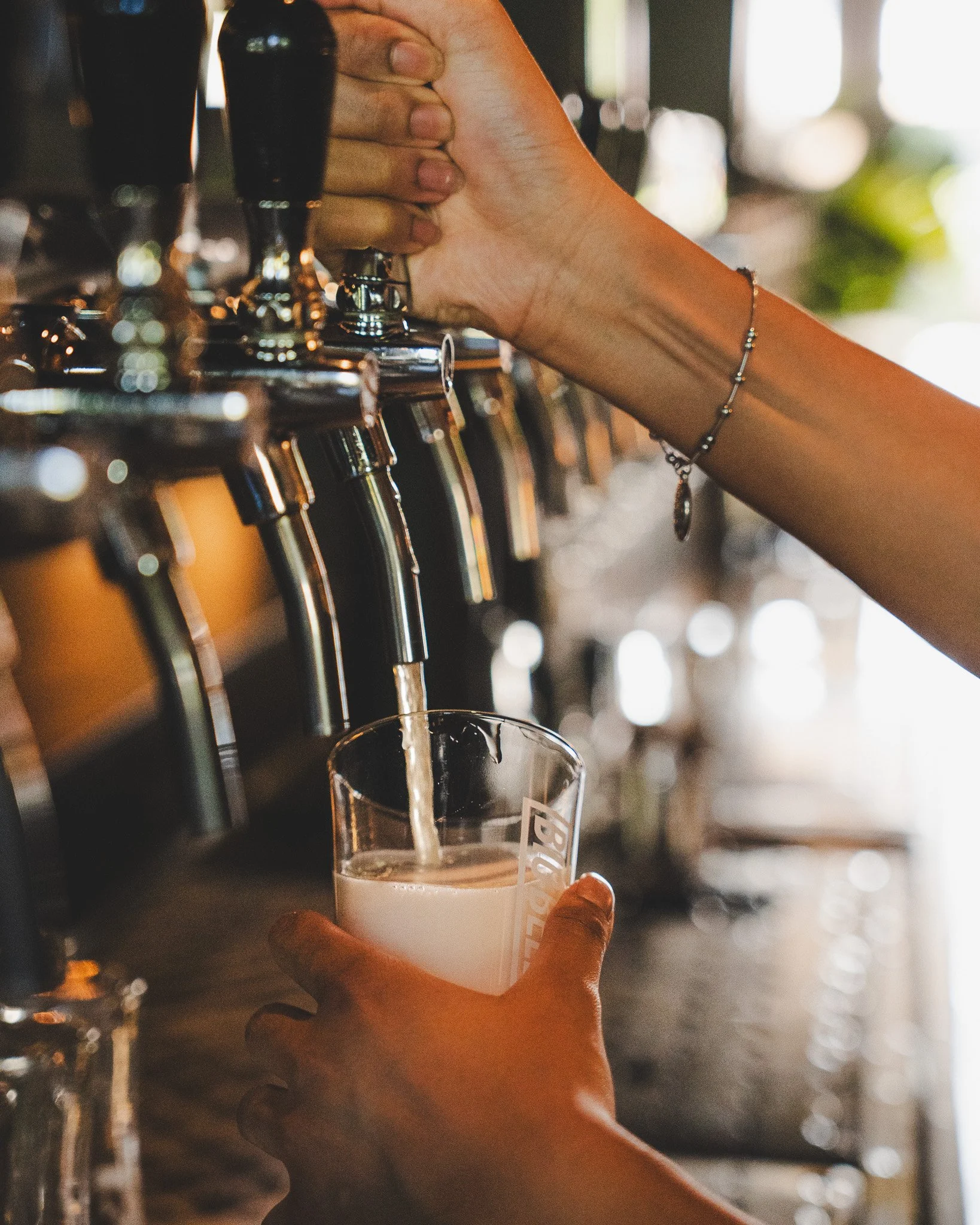 A person holds a clear glass of beer under a tap at a bar, with beer pouring into the glass. The bar has several taps lined up and a blurred background with bright lights.