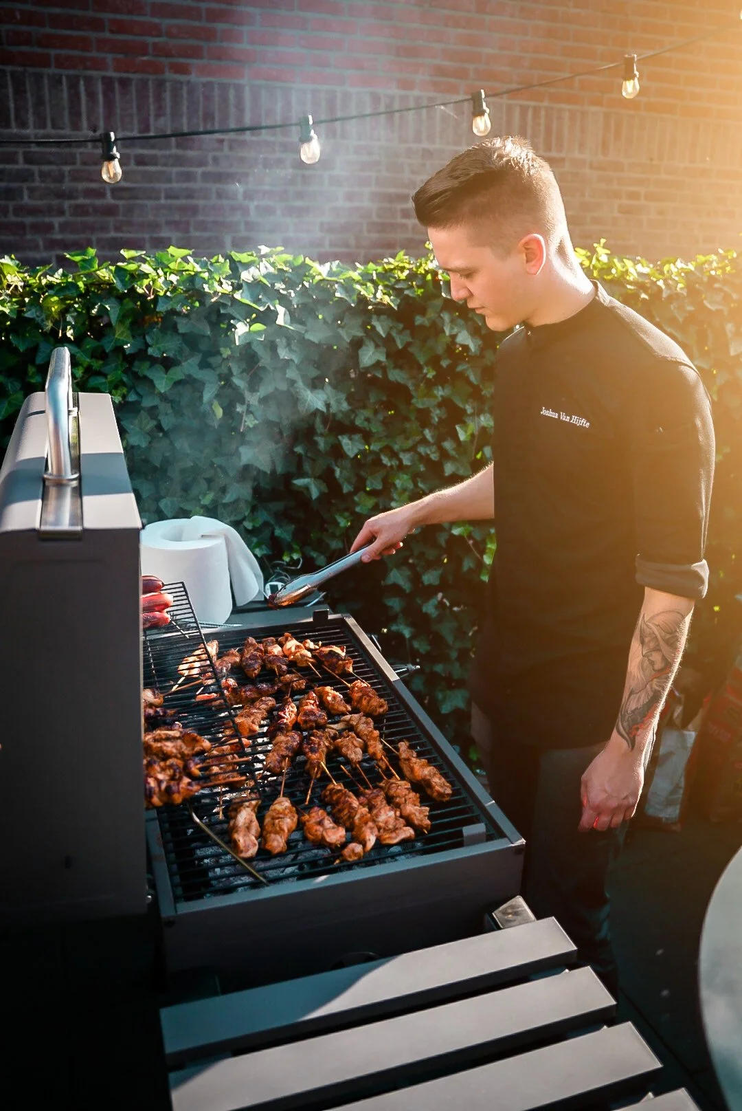 A young man with styled hair, wearing a black t-shirt and black pants, grilling skewered meat outdoors in the evening, with a background of a brick wall, green bushes, string lights, and warm sunlight.