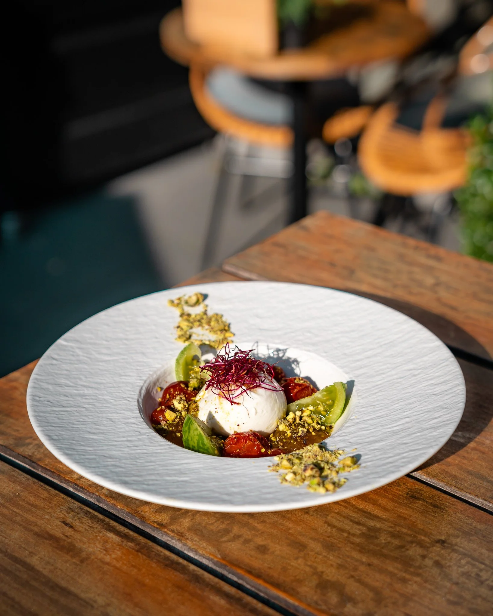 A white plate with a dish of burrata cheese, cherry tomatoes, cucumber slices, and herbs, garnished with crushed pistachios and microgreens, on a wooden table.