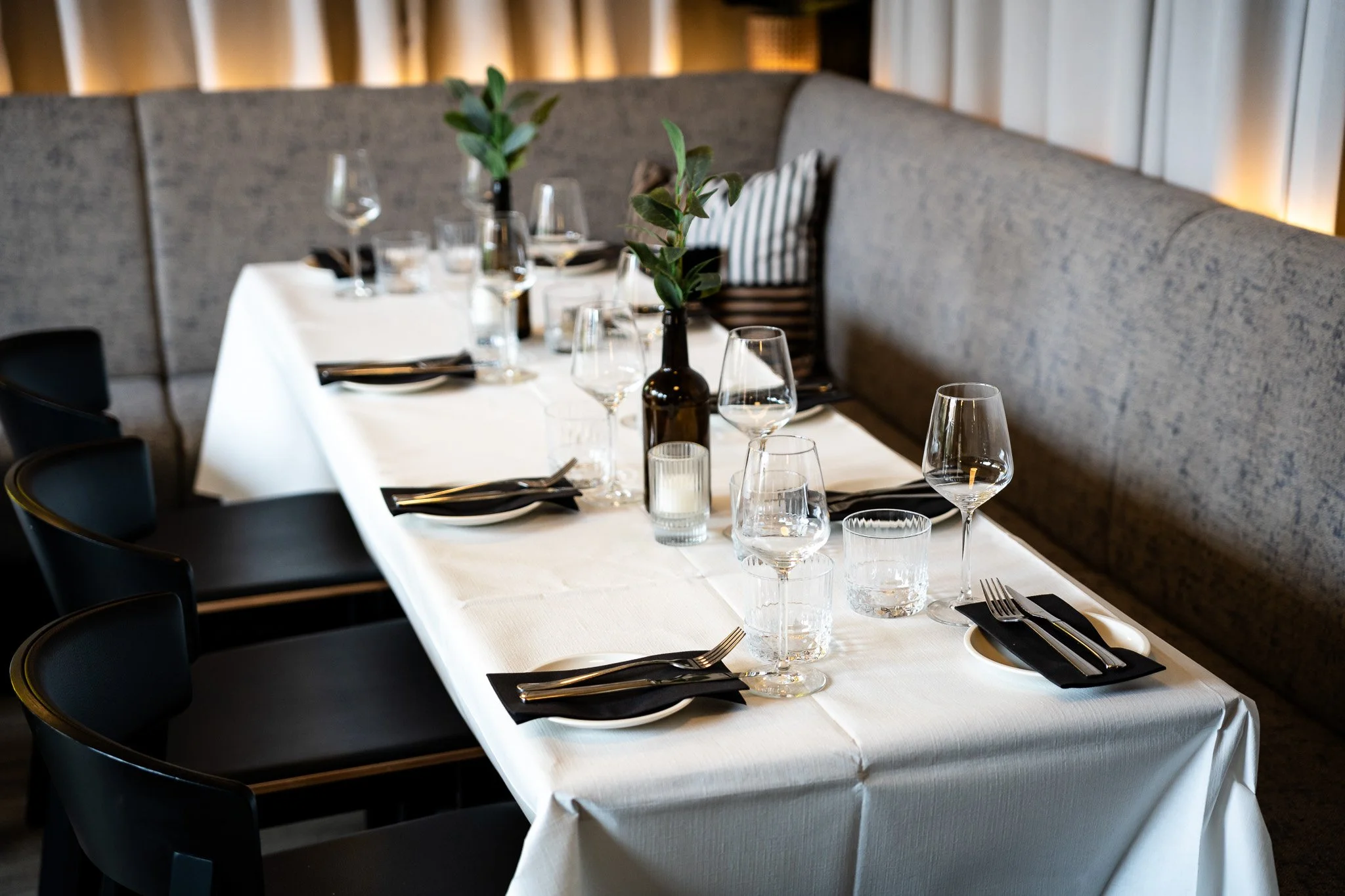 A formal dining table setup with a white tablecloth, multiple wine glasses, water glasses, black napkins, and silverware. A brown bottle with green foliage is placed in the center, and there is a cushioned bench with a striped pillow along the wall. The background features a modern wall design with vertical light accents.