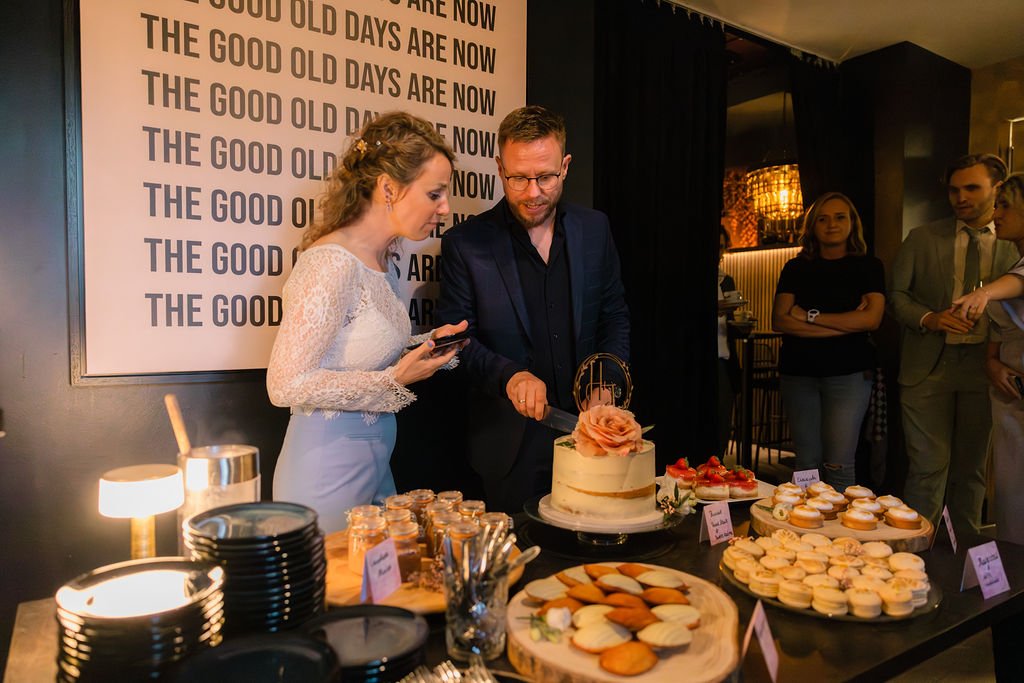 People at a cake table cutting a wedding cake with a floral topper, surrounded by desserts like cookies, cupcakes, and small jars, during a celebration event.