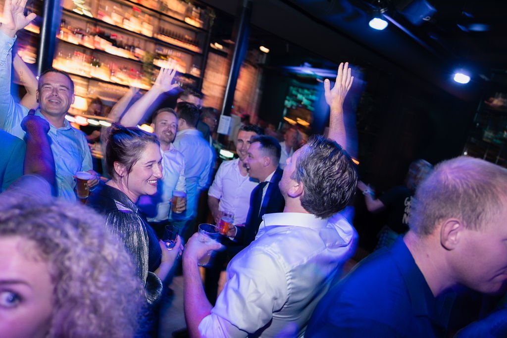 People dancing and socializing in a bar or nightclub, holding drinks, with colorful lighting and shelves of bottles in the background.