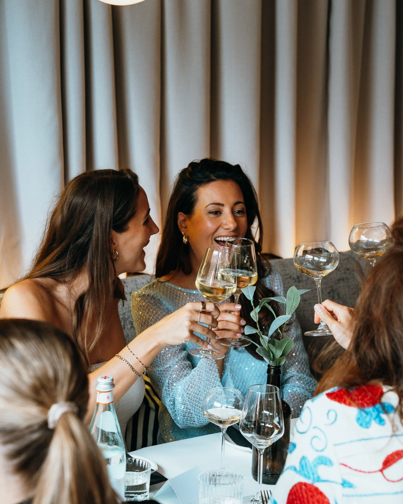 A group of women sitting at a dining table, socializing, and holding glasses of white wine during a celebration or gathering, with a focus on a woman smiling and wearing earrings.