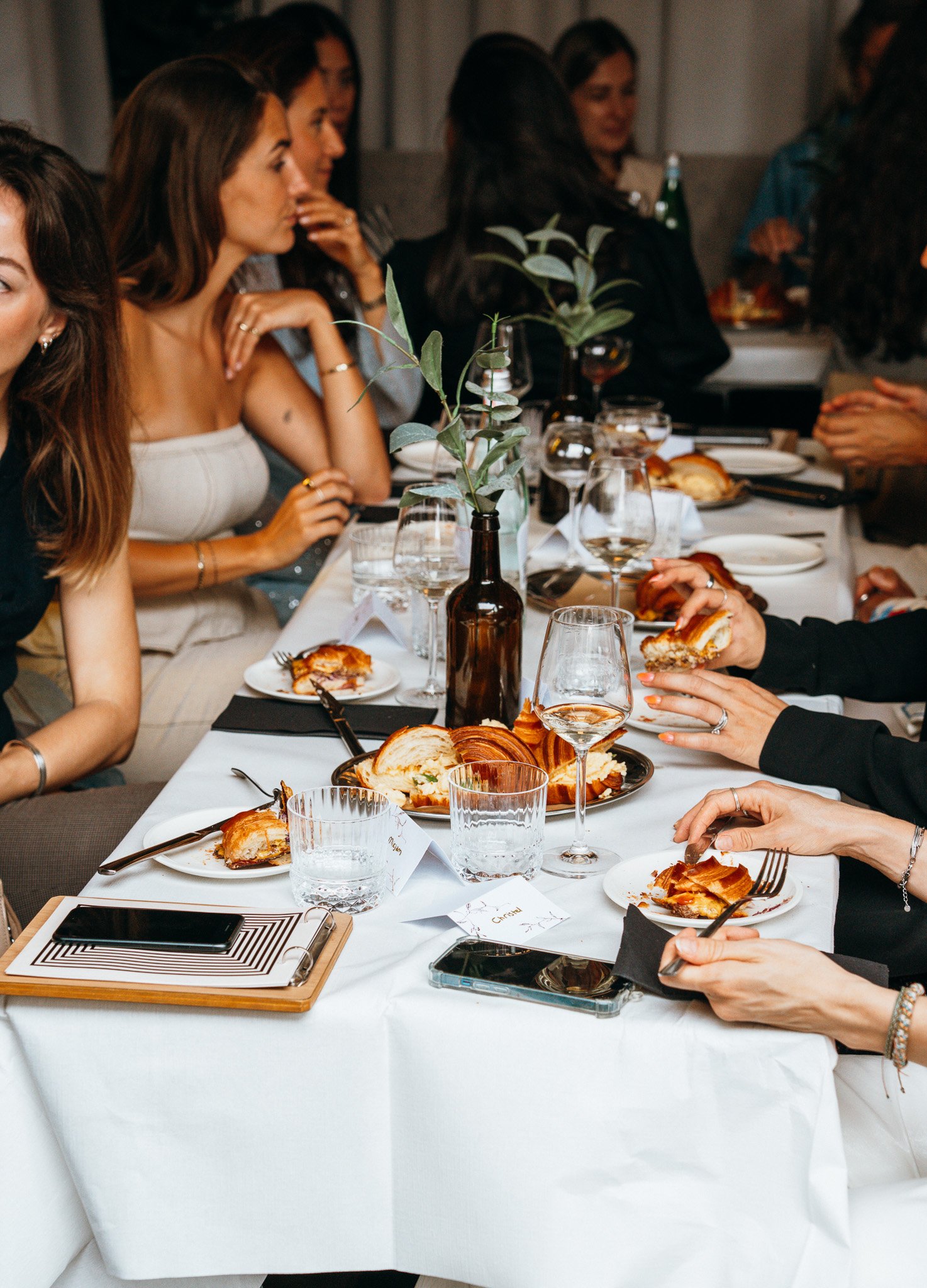 A group of women seated around a dinner table with plates of food, glasses, and cutlery, engaging in conversation during a social gathering.