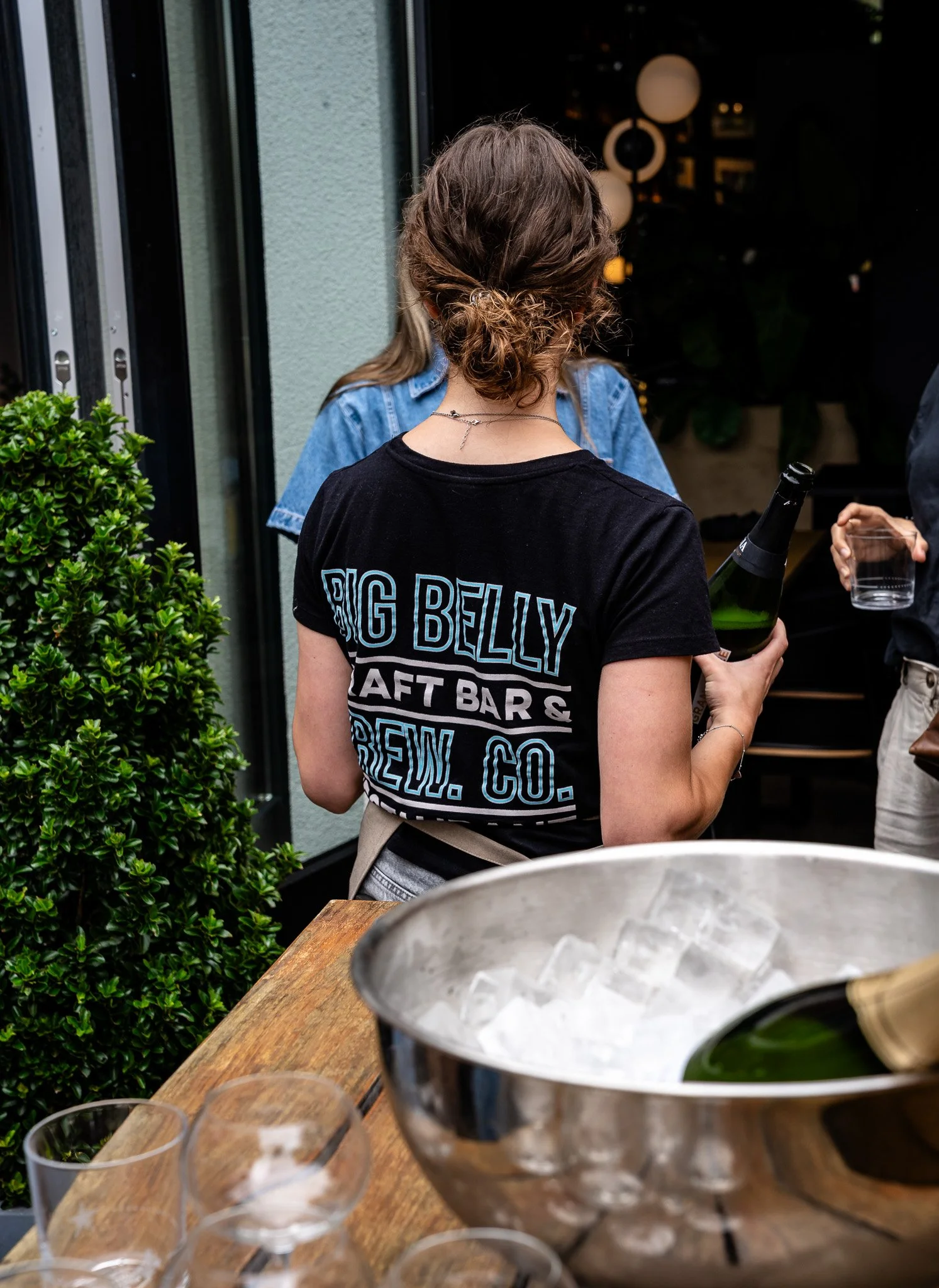 Person with brown hair styled in a messy bun, wearing a black t-shirt that reads "BIG BELLY CRAFT BAR & BREW.CO," holding a bottle of champagne and a clear glass, standing near a gray wall and green shrubbery, at an outdoor gathering with a table and empty glasses.