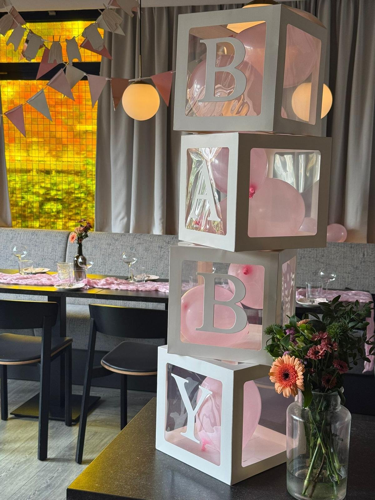 Decorative stacked cubical boxes with pink balloons and cutout letters forming the words "BABY" and "Y" inside the boxes, set up at a decorated indoor event space.