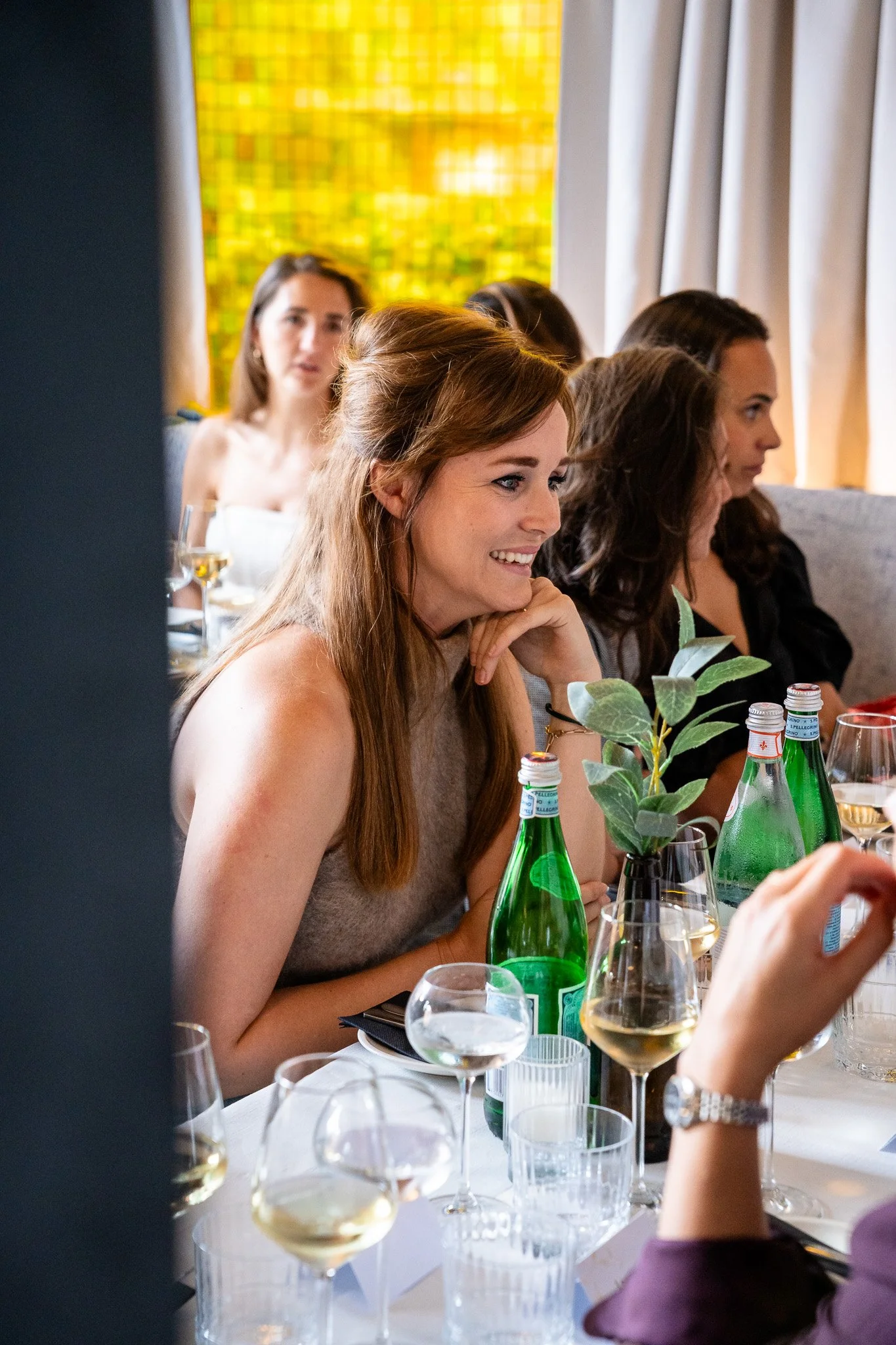 A group of women sitting at a dining table during a gathering or celebration, with wine glasses, bottles of sparkling water, and a small potted plant on the table. Women are smiling and engaged in conversation.