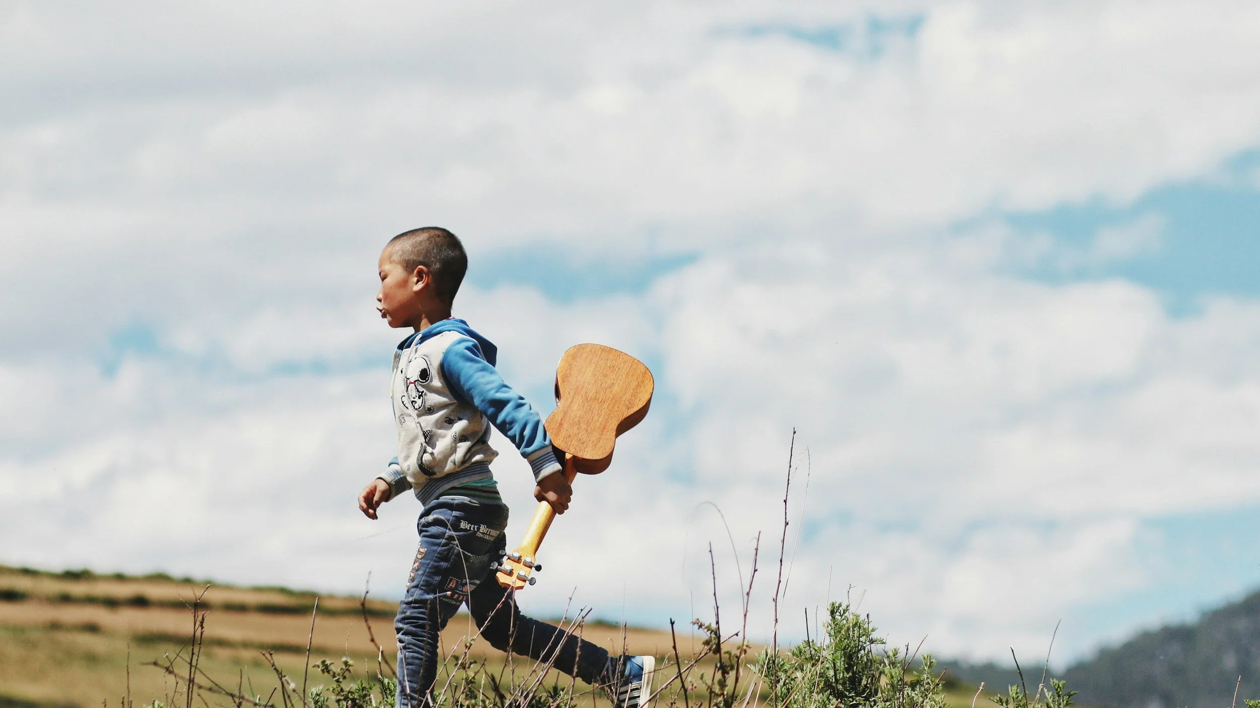 A young boy with a shaved head holding a wooden guitar walking outdoors on a grassy hill against a cloudy sky.