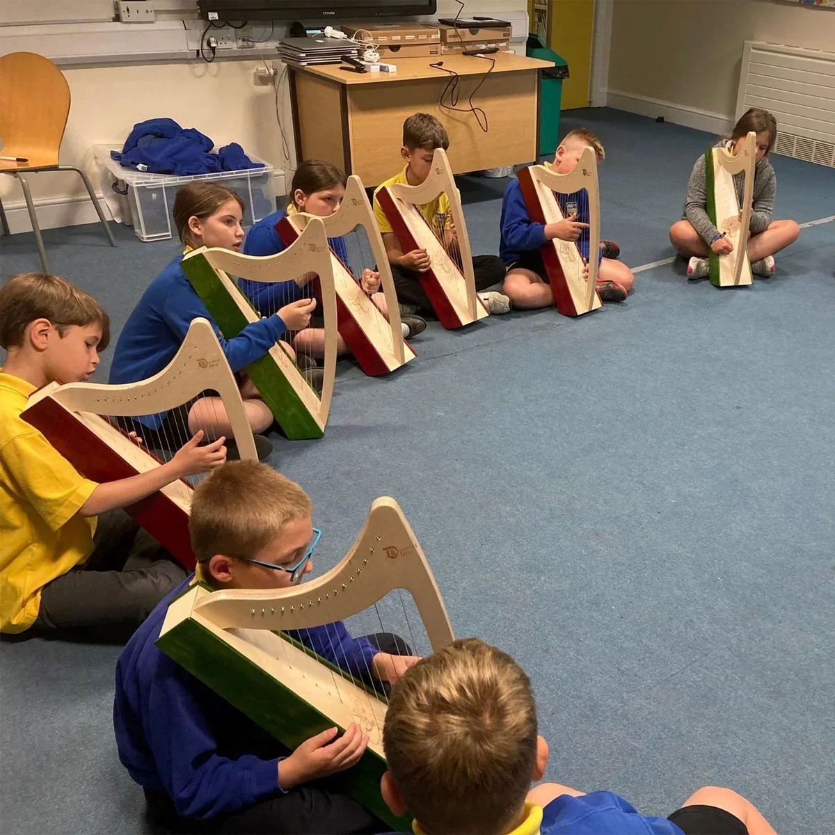 A group of children sitting on the floor, playing upright harps in a classroom setting.
