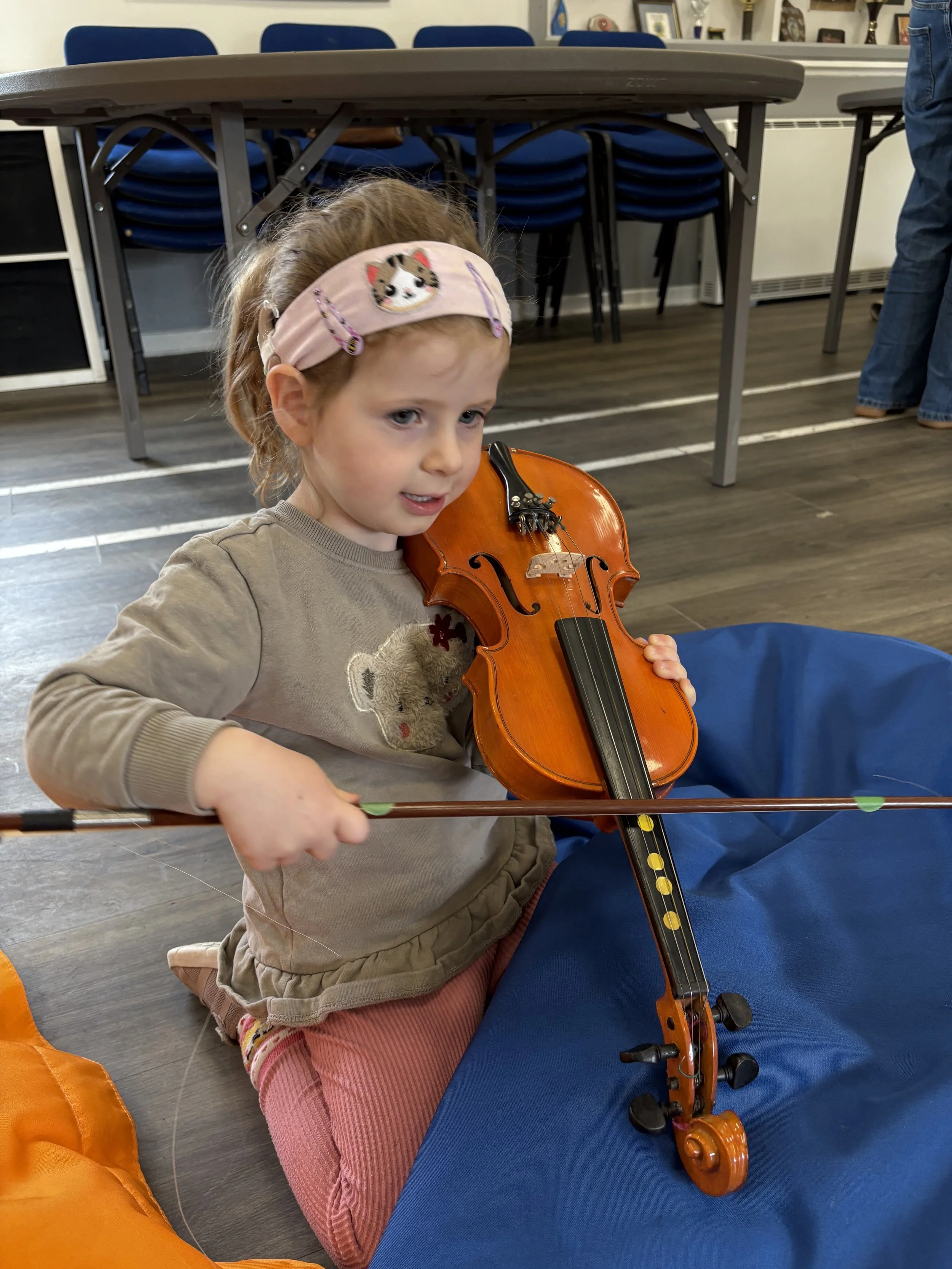 A young deaf girl with light brown hair, wearing a headband with dog design, is sitting on the floor playing a small wooden violin. She is looking down at the violin while holding the bow in her right hand.