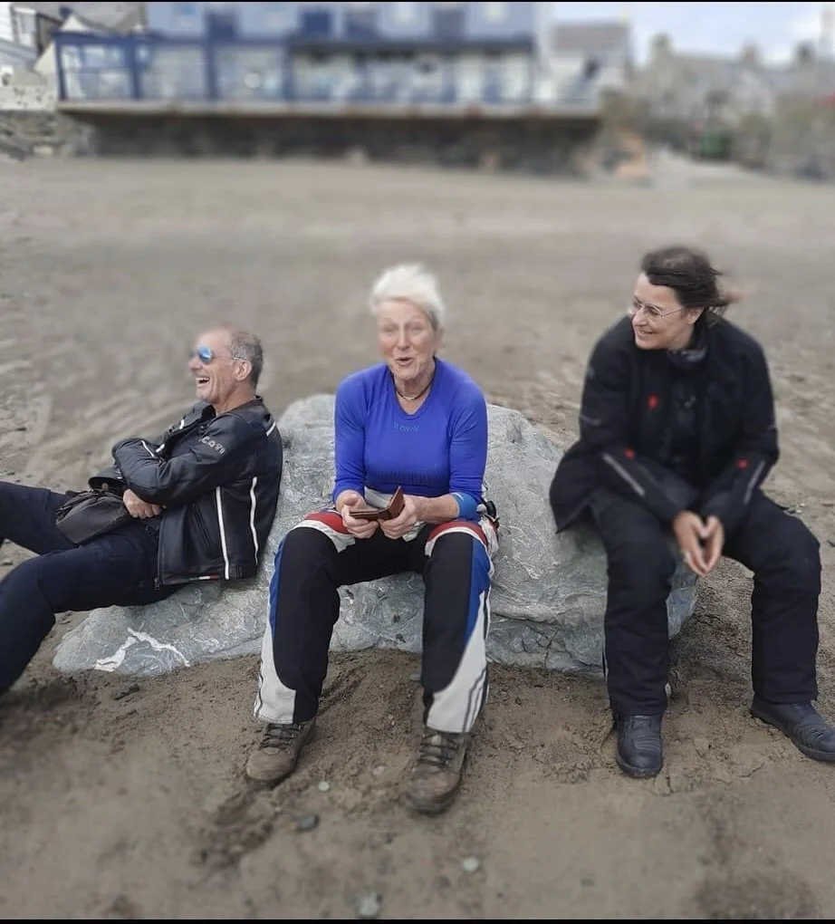 Three people sitting on a large rock on a beach, smiling and laughing. The person on the left is a man wearing sunglasses and a black jacket. The person in the middle is a woman with short white hair, wearing a blue shirt and black and white pants, holding a wallet. The person on the right is a woman with brown hair, wearing a black jacket and black pants.