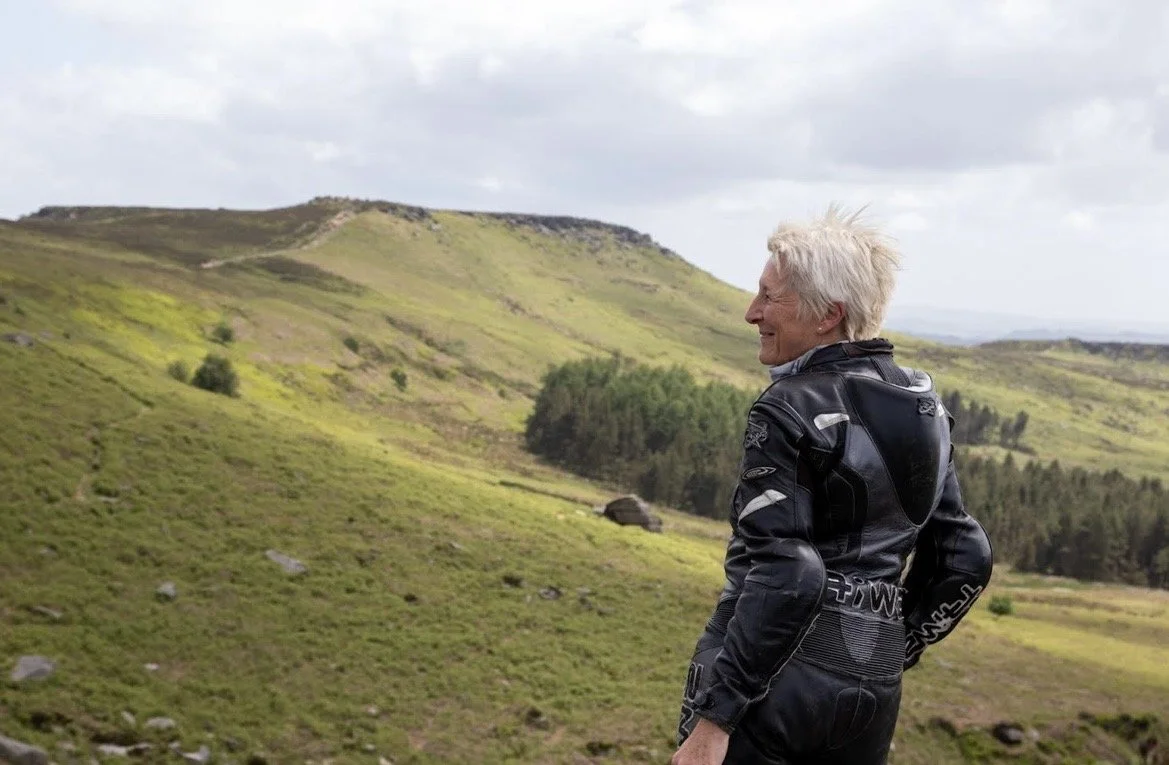 Older woman in black motorcycle jacket standing outdoors in a green hilly landscape under cloudy sky.