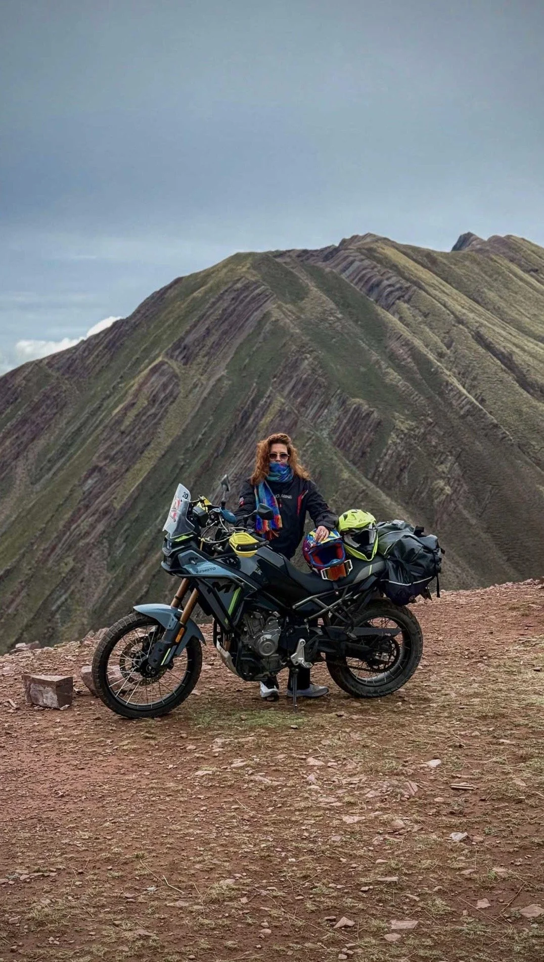 A woman with curly hair and sunglasses standing next to a black motorcycle with luggage, helmets, and gear, on a dirt path with mountains in the background.
